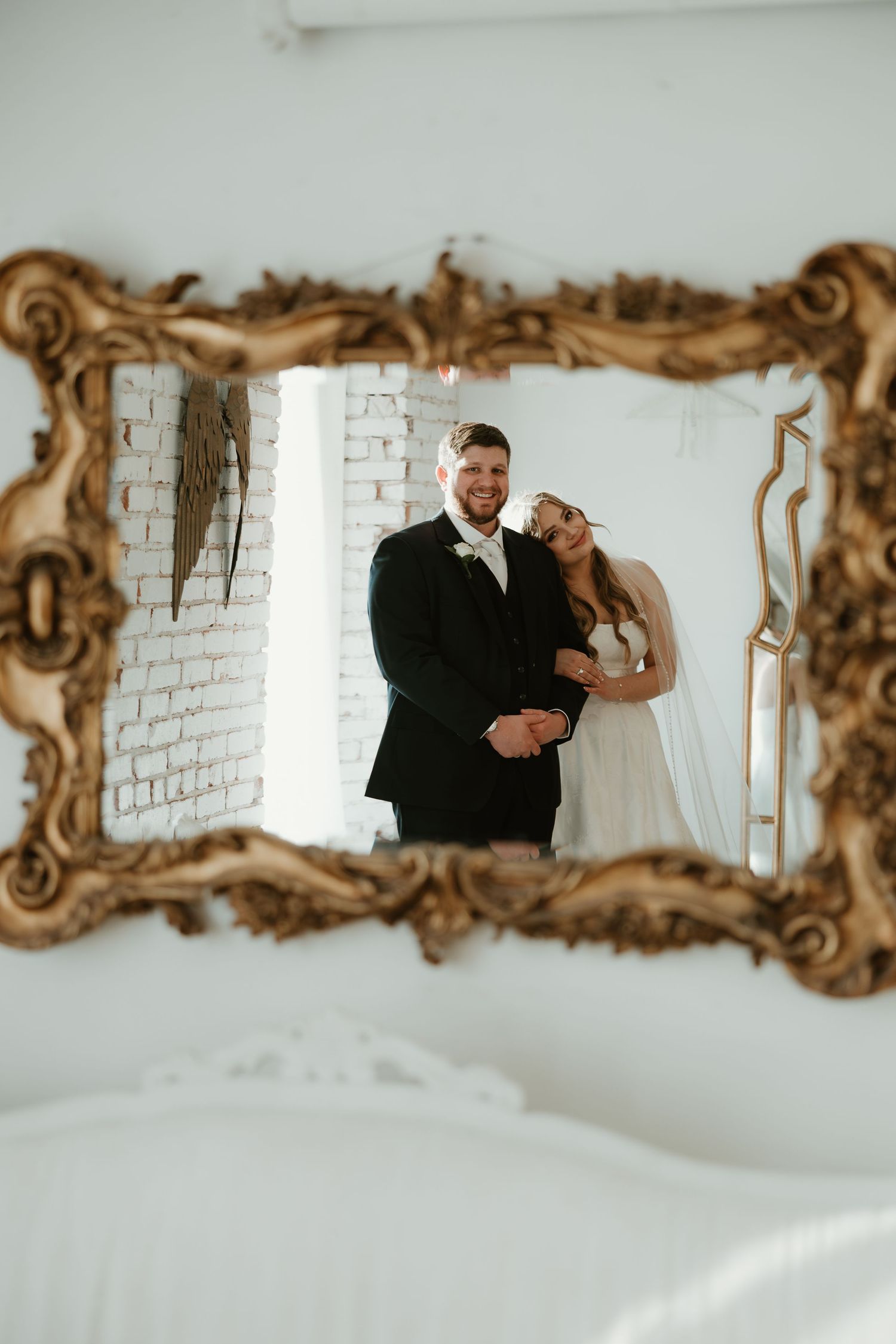 Wedding couple's reflection captured in an ornate gold vintage mirror against a white brick wall background.
