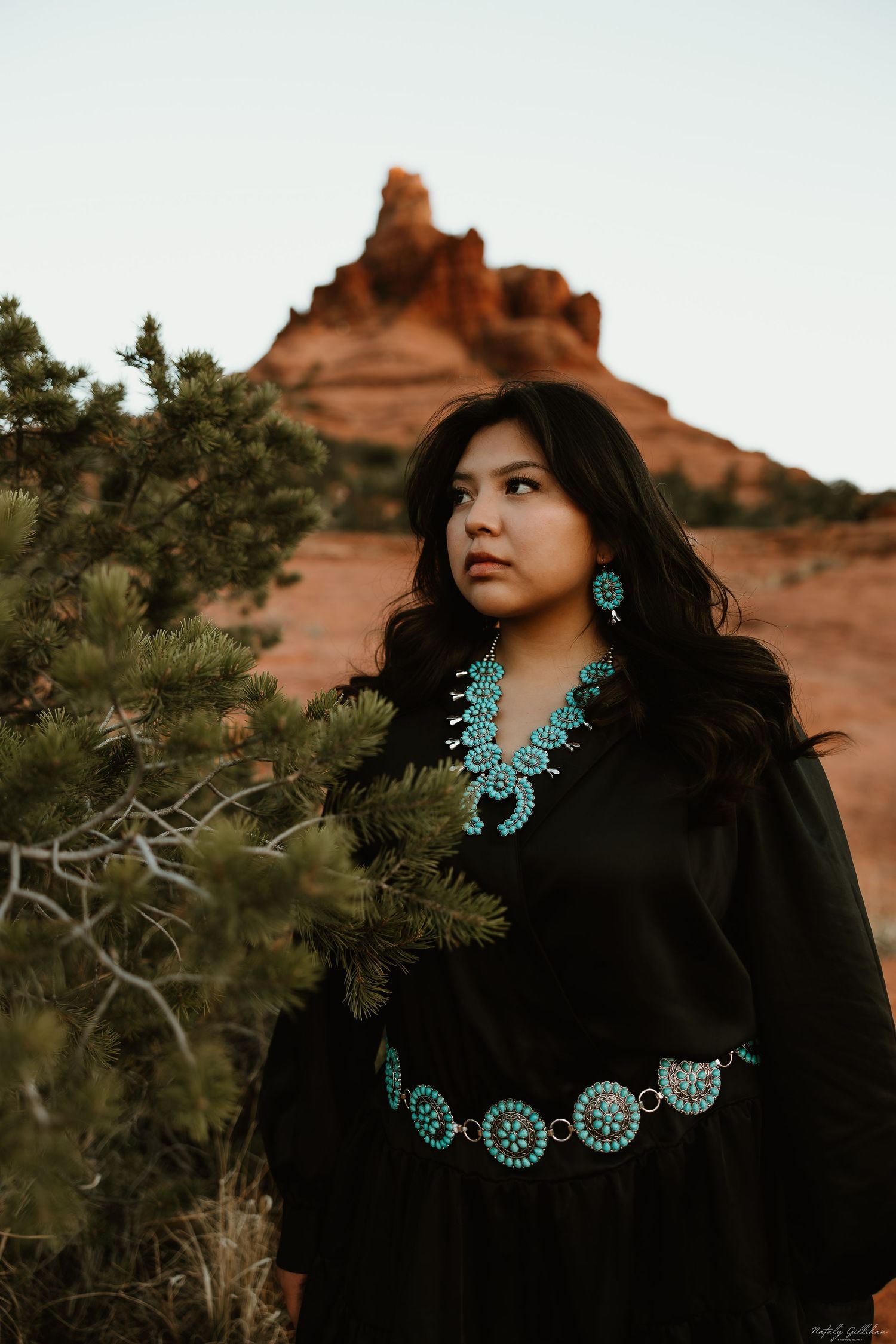 Traditional turquoise jewelry adorns a black dress with red desert rocks and desert foliage in the background at dusk.