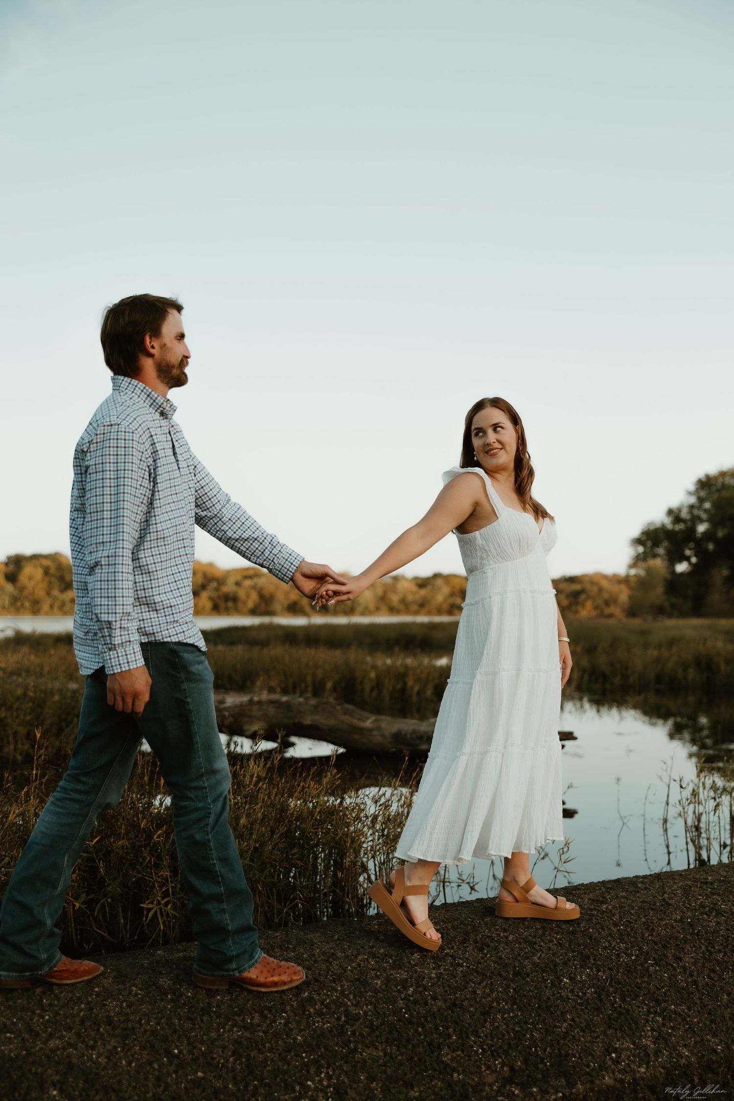 Couple shares a romantic moment near a lake at sunset, with the woman in a flowing white dress.