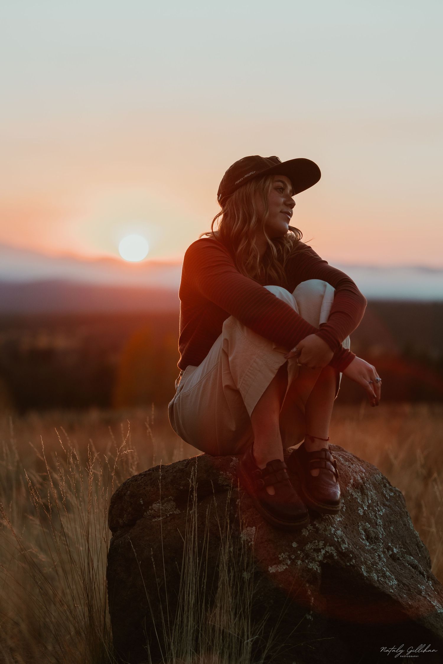A silhouetted figure in a baseball cap sits on a rock watching the golden sunset over mountains and dry grass fields.