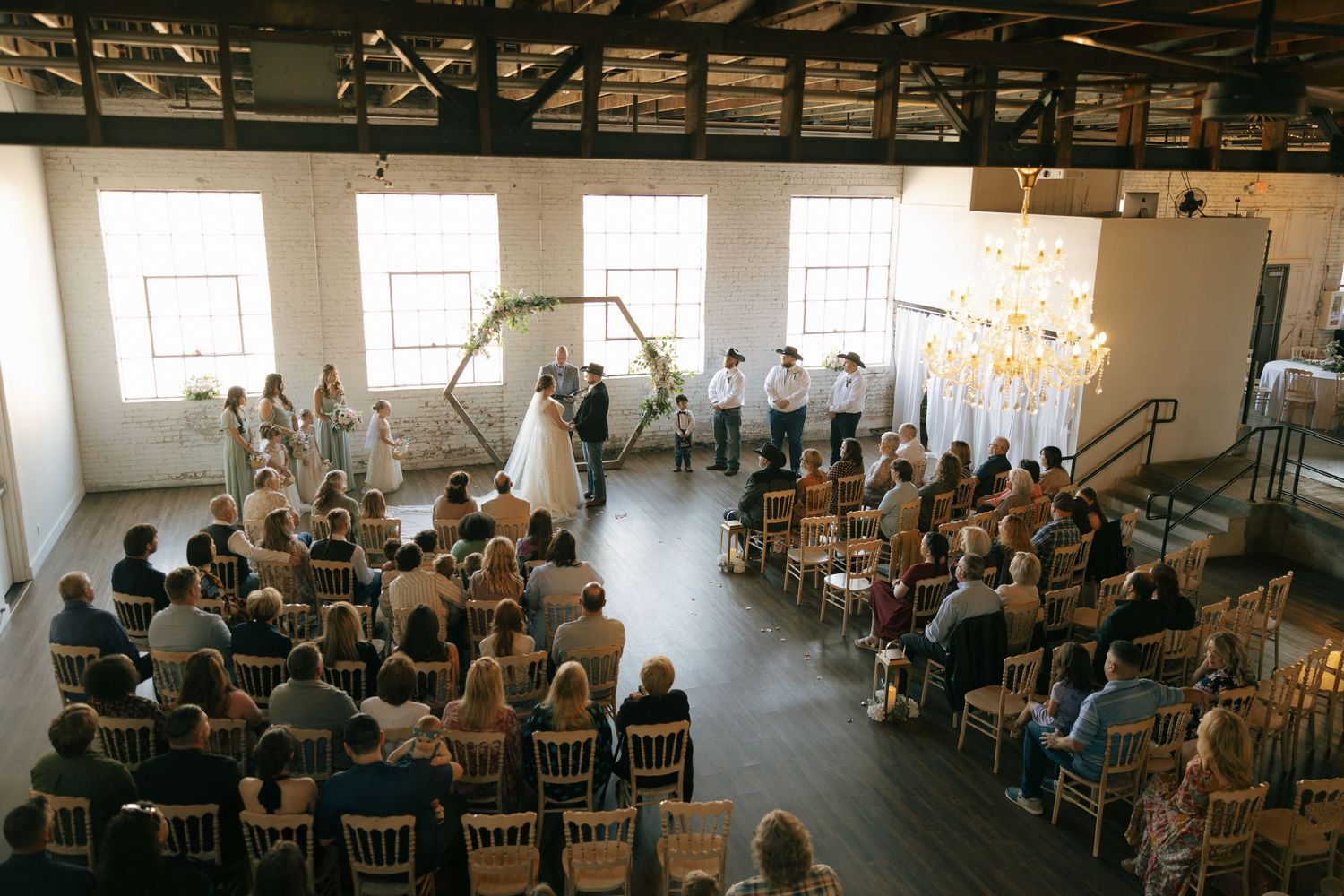 Aerial view of an elegant indoor wedding ceremony with guests seated in rows facing a floral arch altar in a bright loft venue.