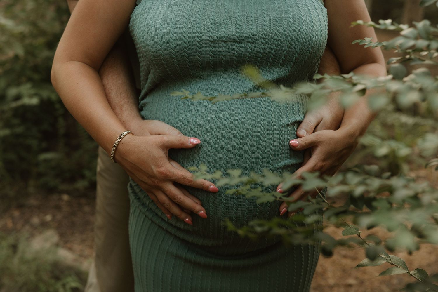 A person in a green sweater lovingly cradles their pregnant belly in a nature setting with soft sunlight filtering through.