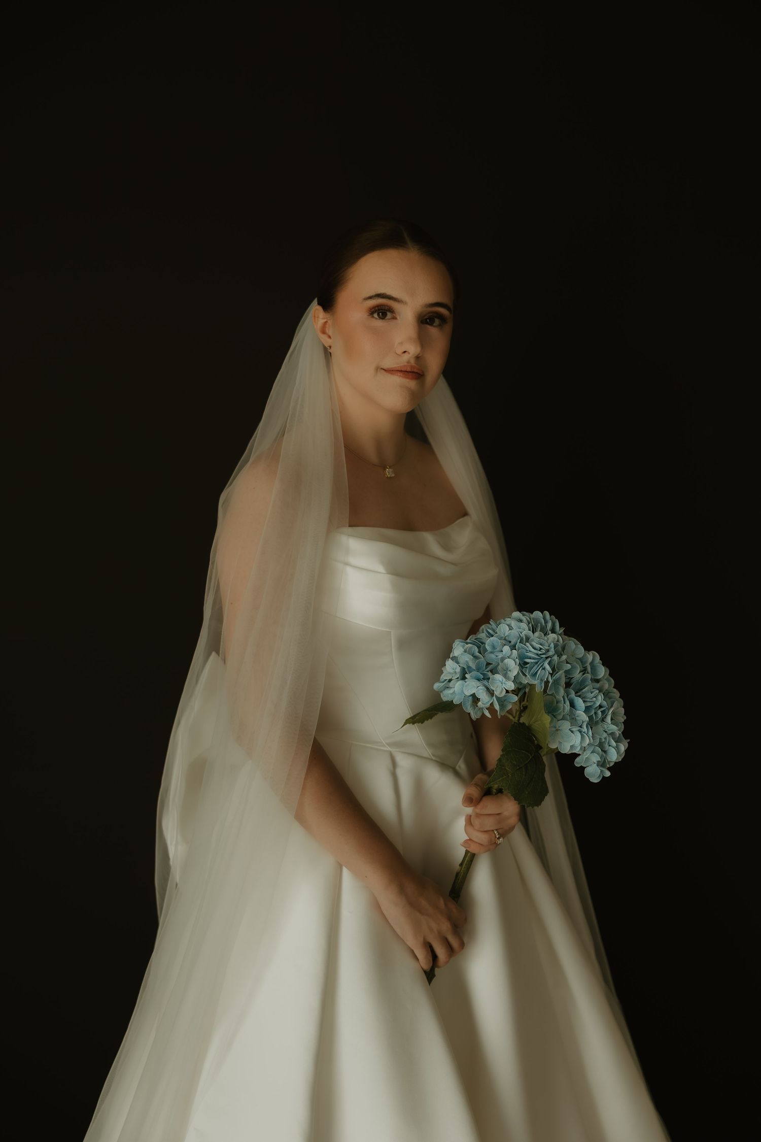 Bride in white satin gown and veil holds blue hydrangea bouquet against dark background.