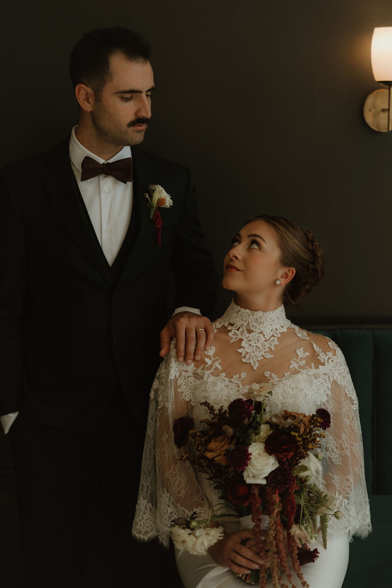 Bride in lace cape gown holds burgundy bouquet beside groom in black tuxedo with bow tie in moody wedding portrait.