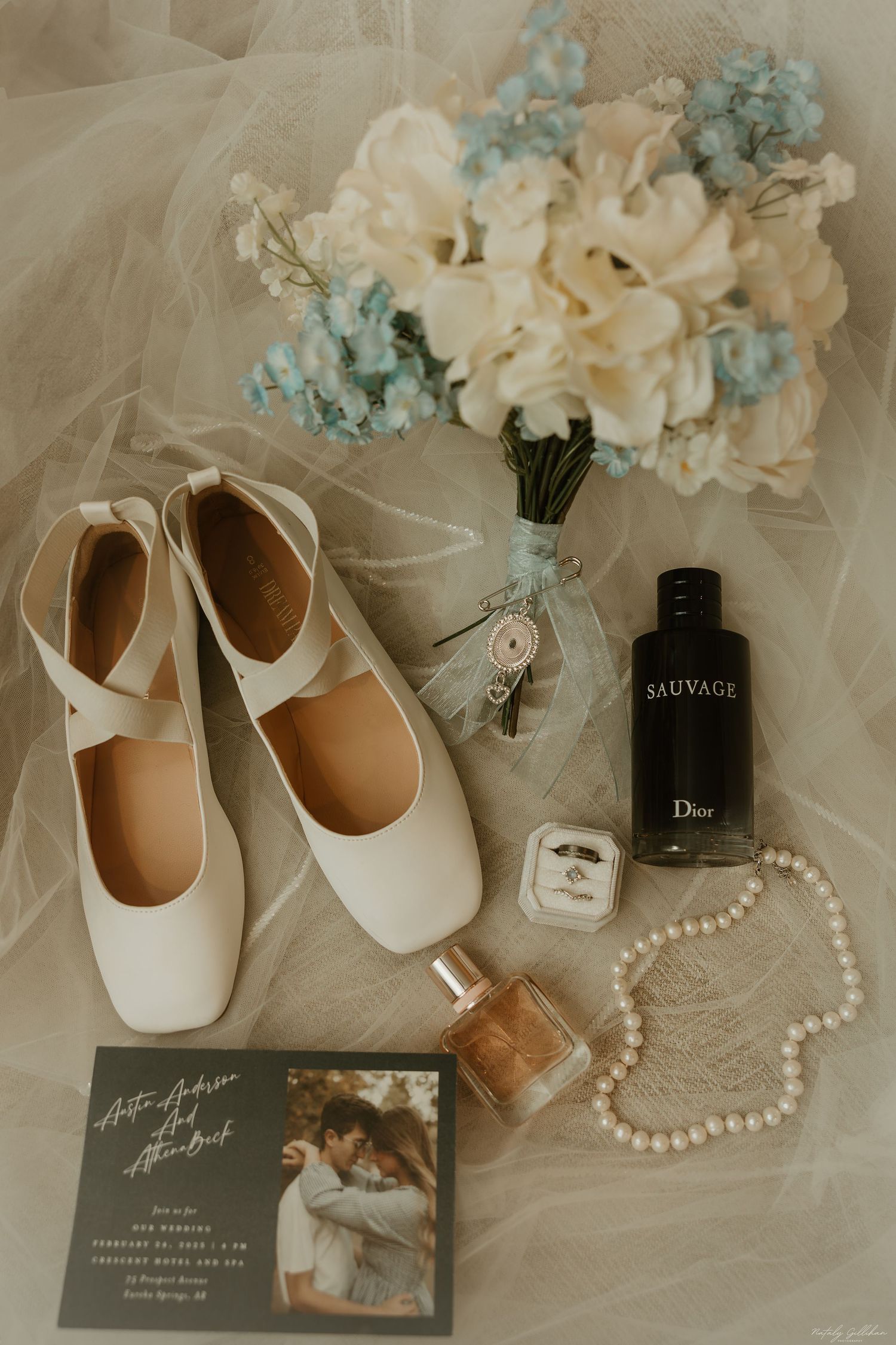 Wedding accessories including white shoes, flowers, perfume and jewelry arranged on tulle fabric.