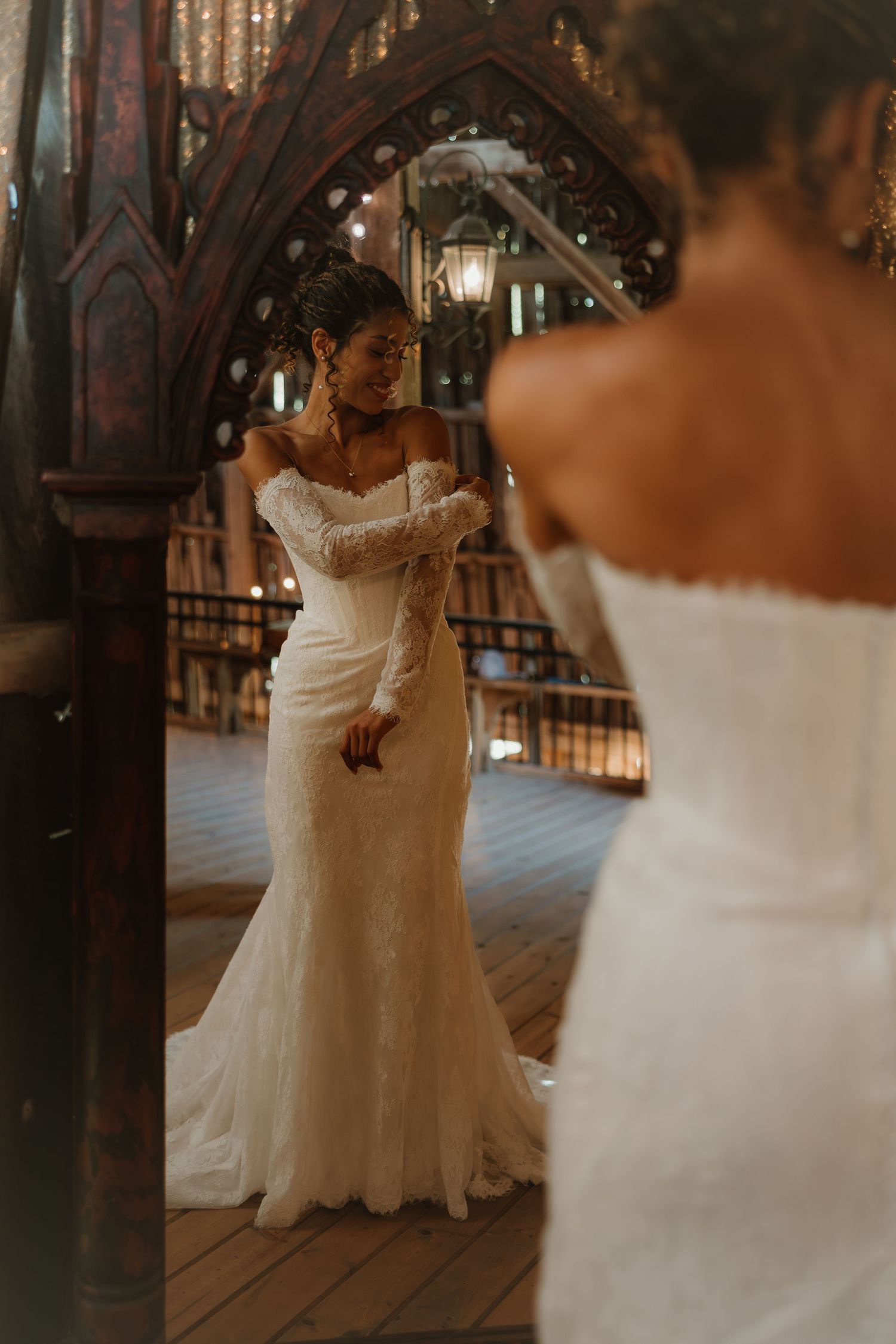 A bride in an elegant off-shoulder mermaid wedding gown admires her reflection in an ornate gothic-style mirror.