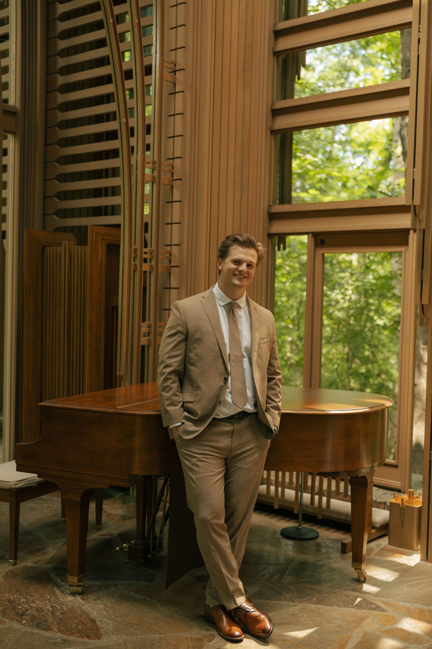 Young man in tan suit leaning against grand piano in elegant wood-paneled room with floor-to-ceiling windows.