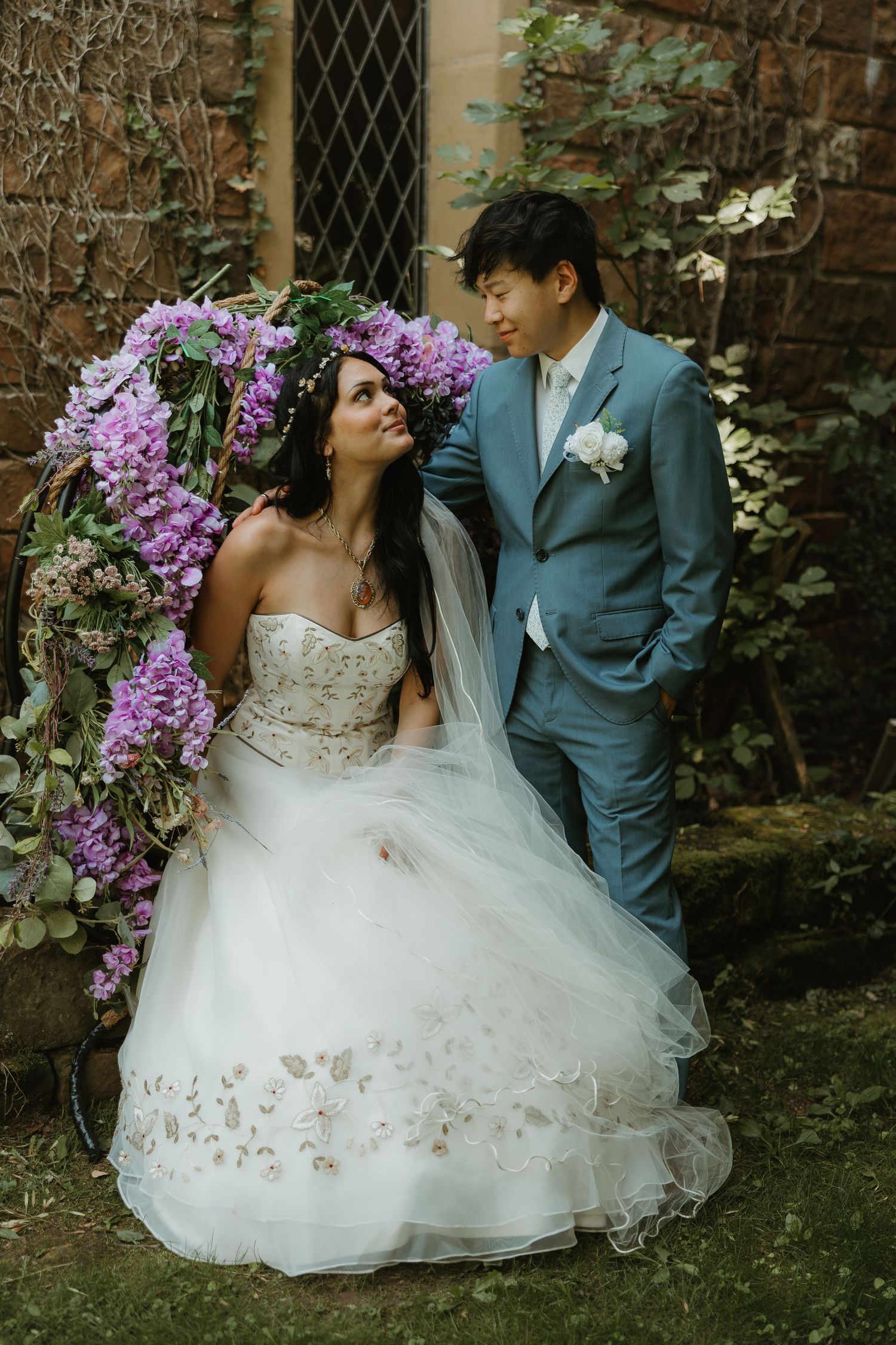 A romantic wedding photo series showing a couple in formal attire with purple floral decorations against a stone wall background.