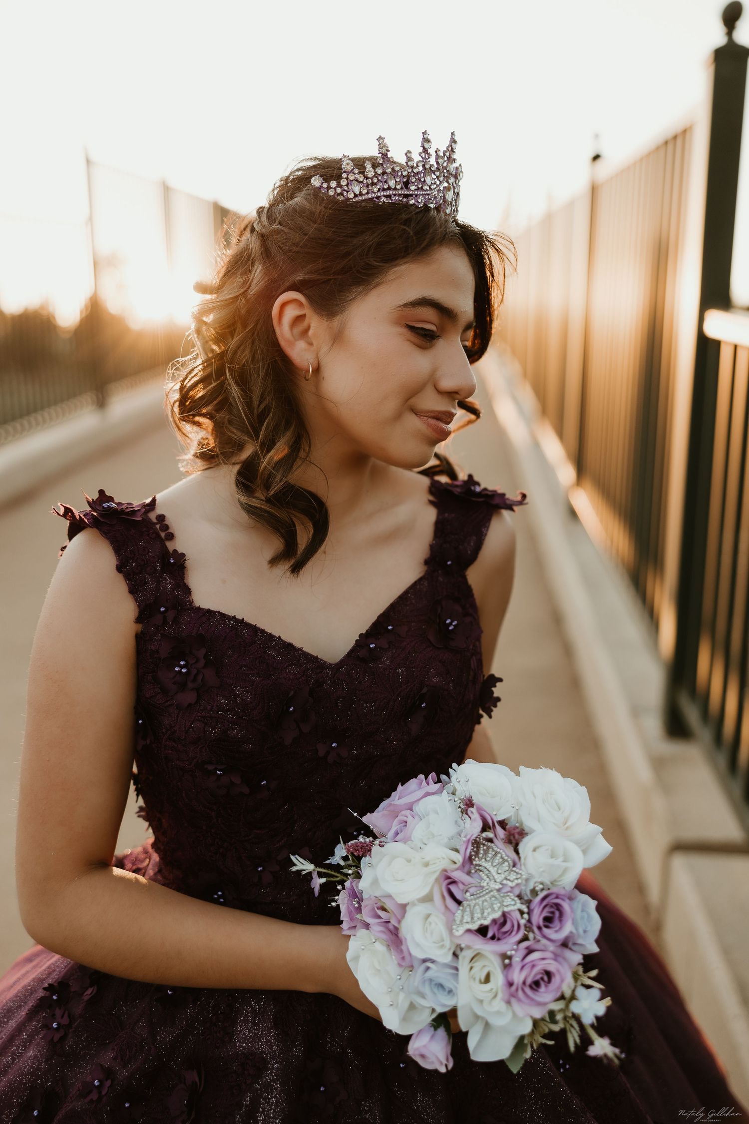 A young woman in a deep purple gown holds a pink and white bouquet while wearing a tiara at sunset.