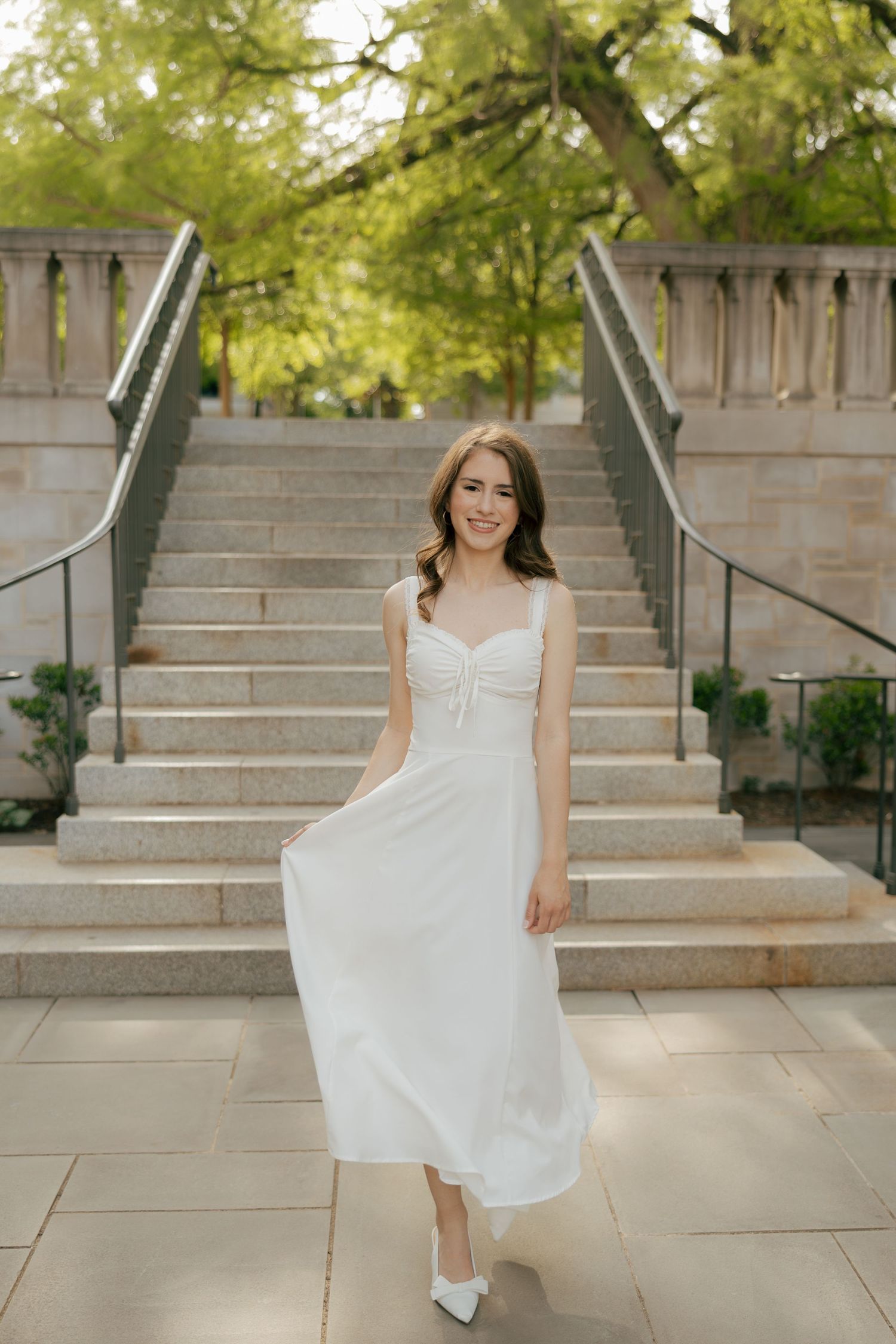 Young woman in white midi dress and heels poses gracefully on stone steps surrounded by lush green trees.