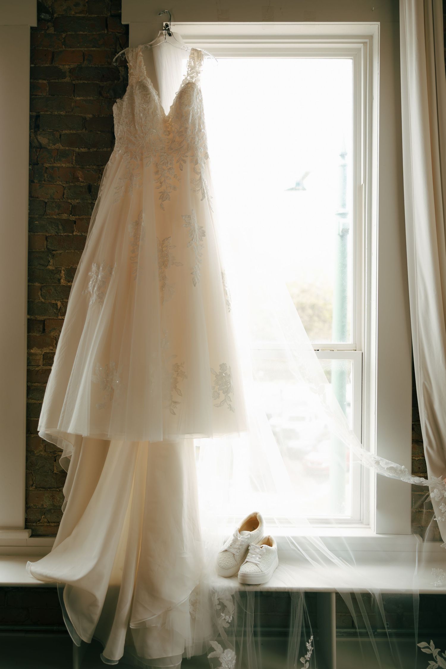 Lace A-line wedding dress hanging in window light with white sneakers and veil on windowsill below.