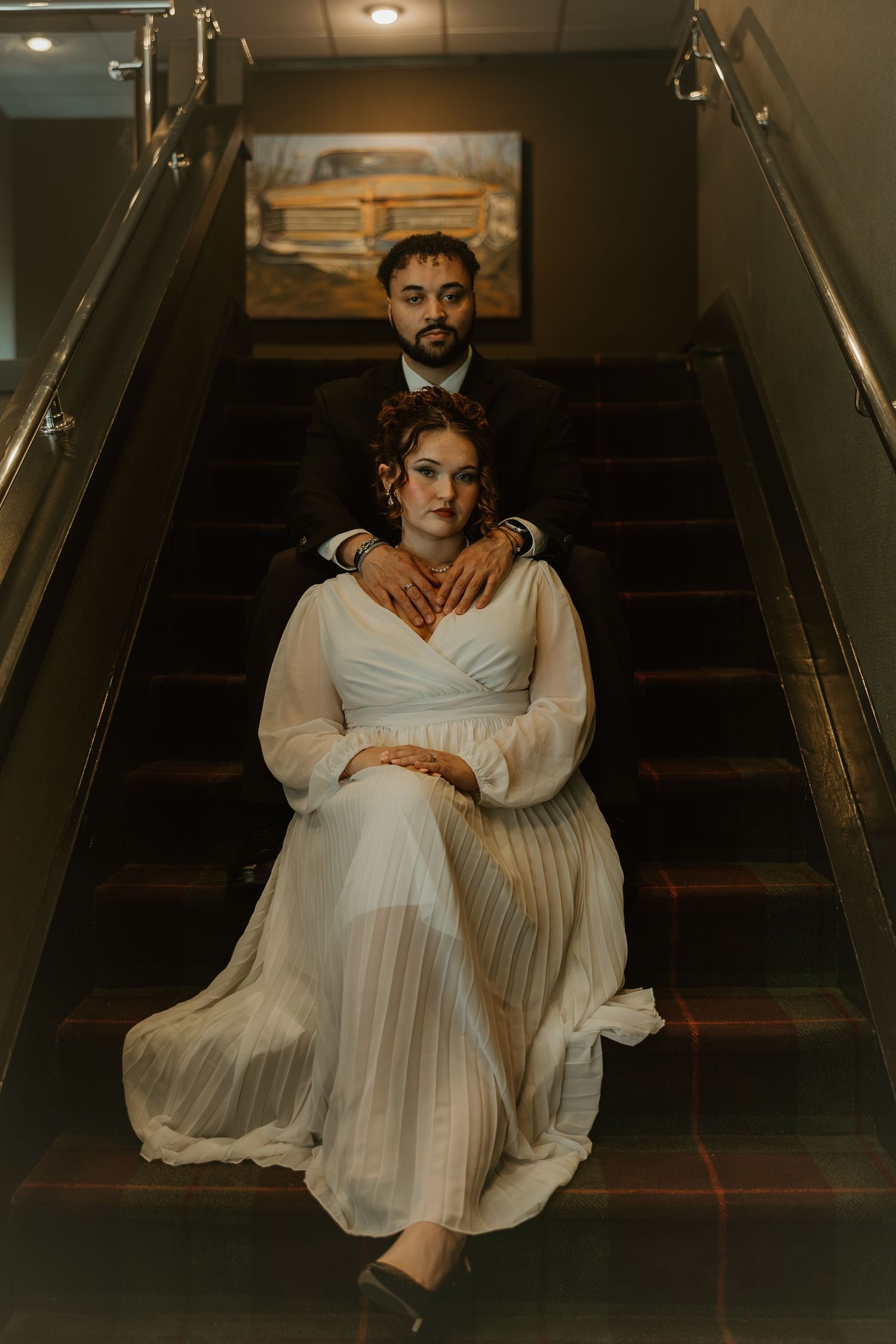 A person in a flowing white dress poses dramatically on dark wooden stairs in moody lighting.