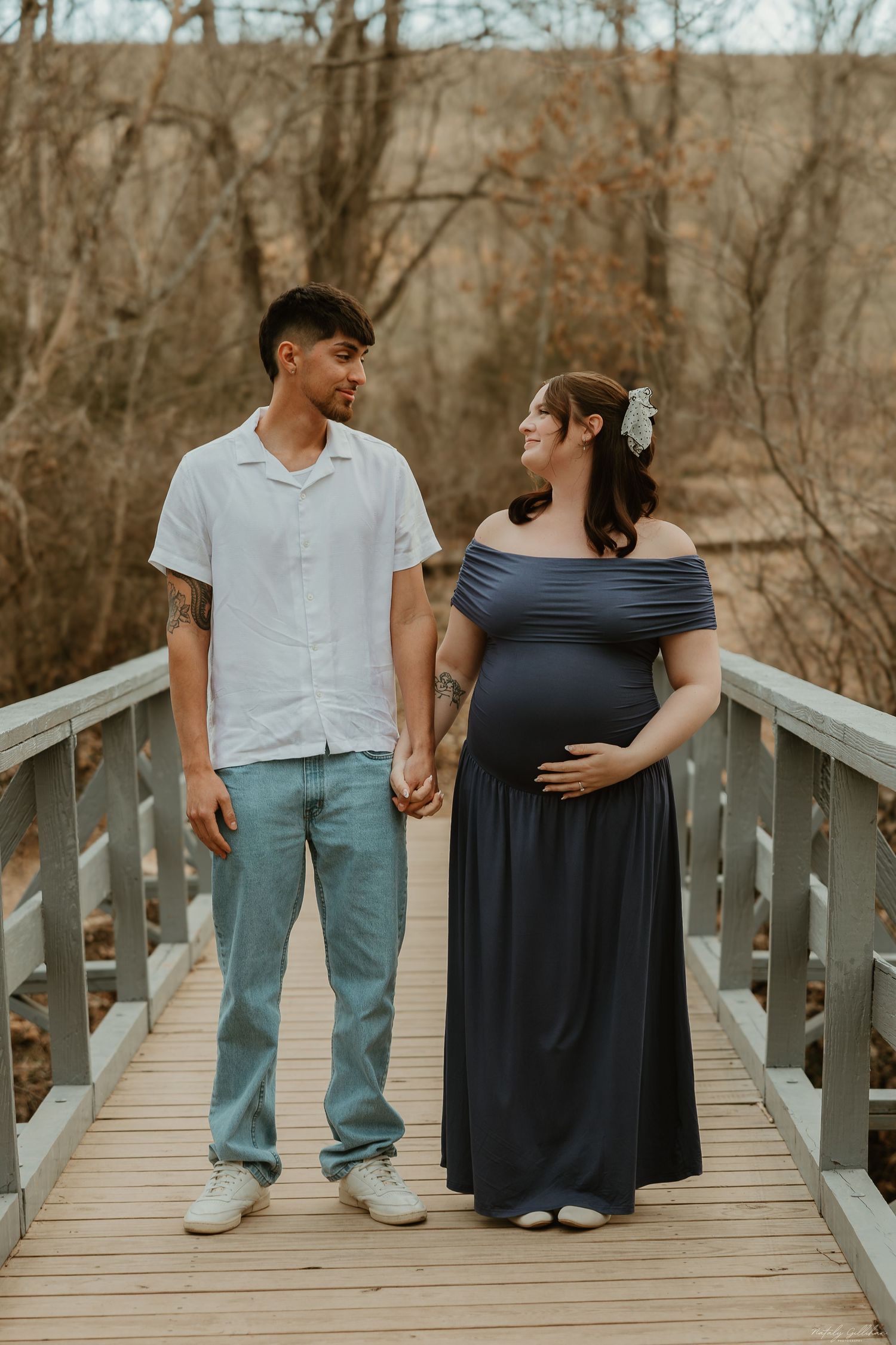 Expectant couple holding hands while standing on a wooden bridge in autumn surrounded by bare trees.