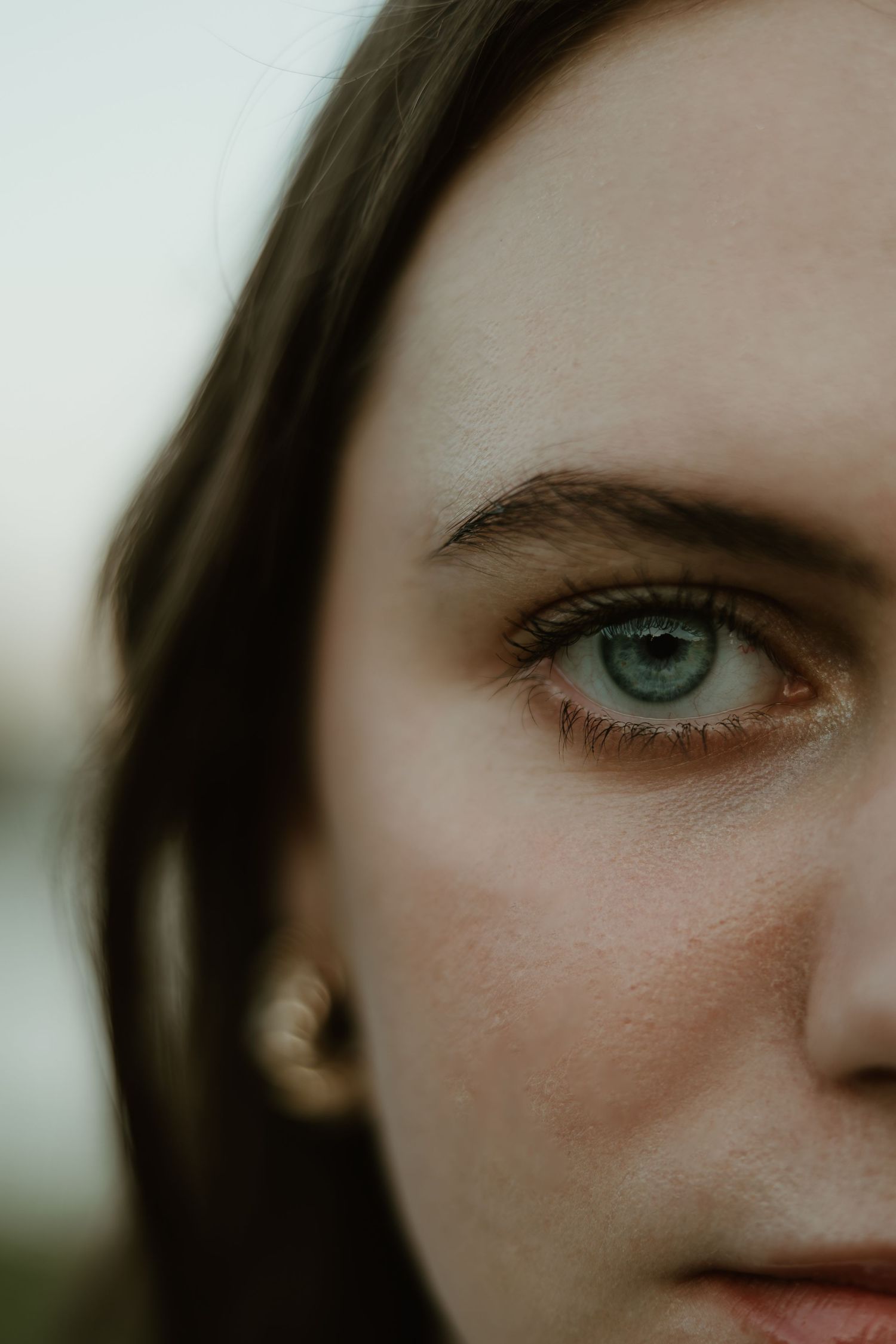 Close-up portrait showing half of a face with striking blue-green eyes and dark hair against a light background.