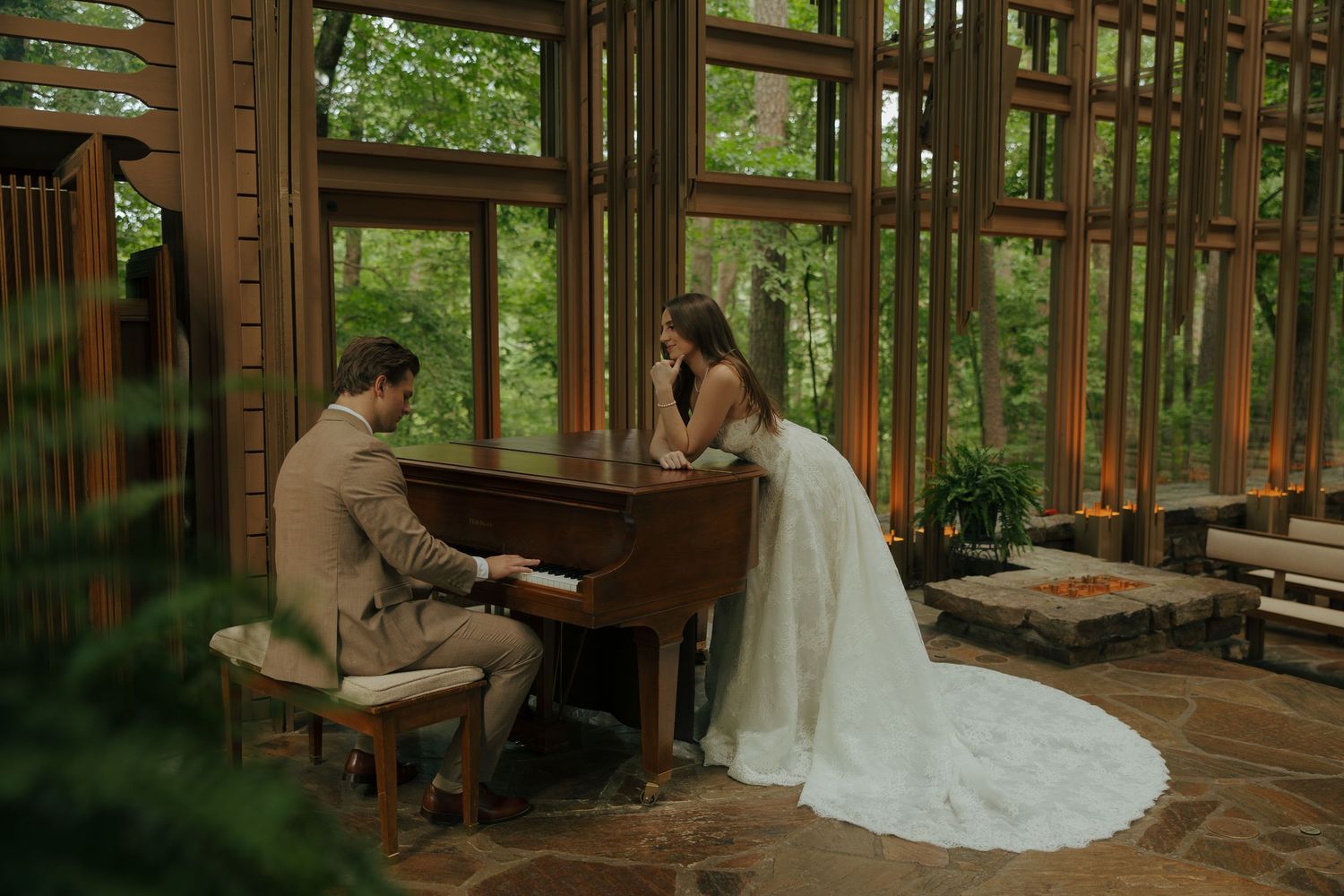Groom playing piano for bride in elegant wood-paneled chapel surrounded by lush green trees on their wedding day.