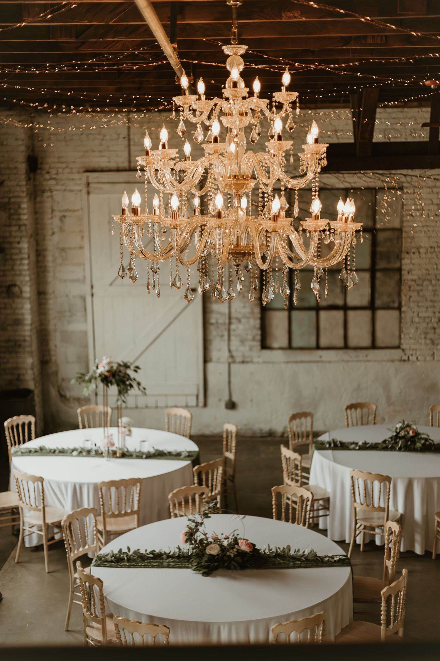 Elegant wedding reception in rustic barn venue with crystal chandelier, round tables with white linens and greenery centerpieces.