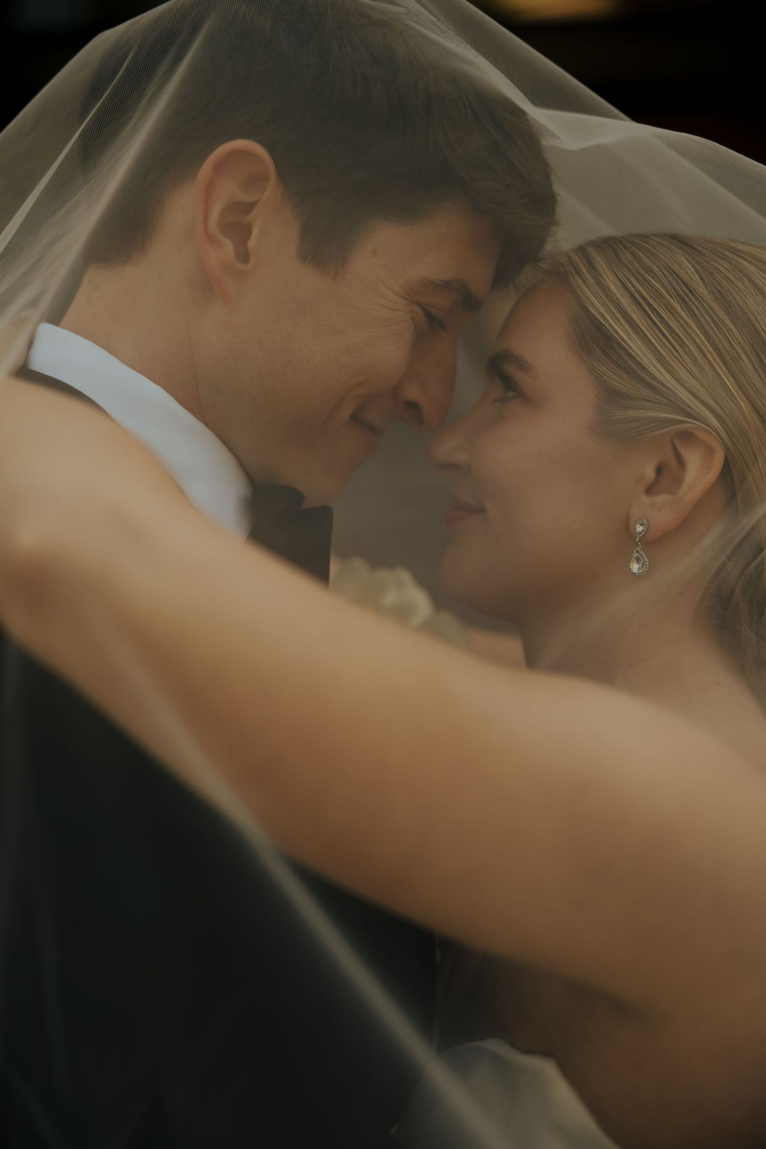 A bride and groom share a romantic moment under a wedding veil, smiling at each other in a tender embrace.
