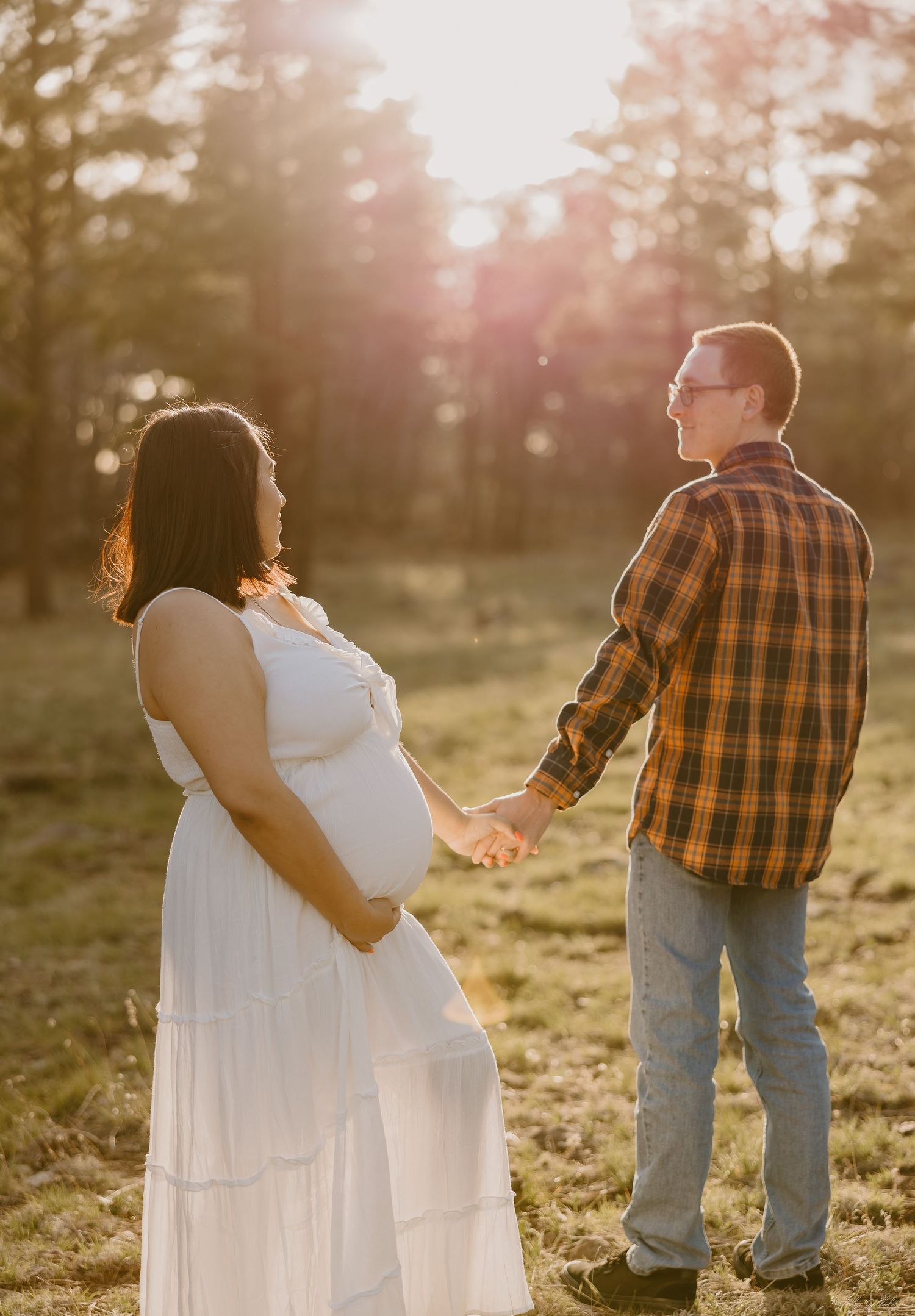 Couple holding hands during sunset maternity photo session in field with golden sunlight glowing through trees.