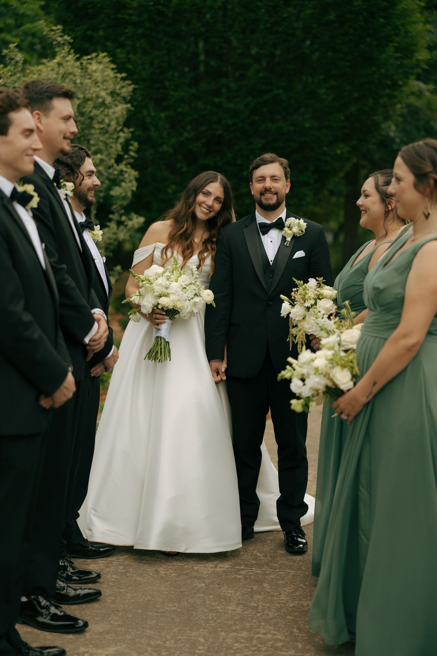 Bride and groom hold hands smiling with wedding party in green dresses and black tuxedos outdoors.
