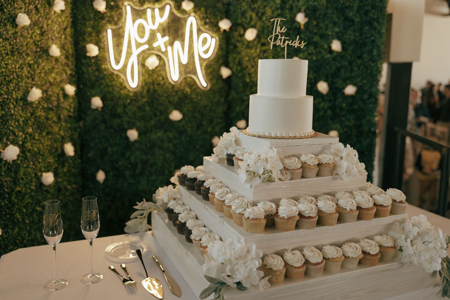 Elegant wedding cake display with cupcake tower, white floral decor, and a glowing You & Me neon sign backdrop.