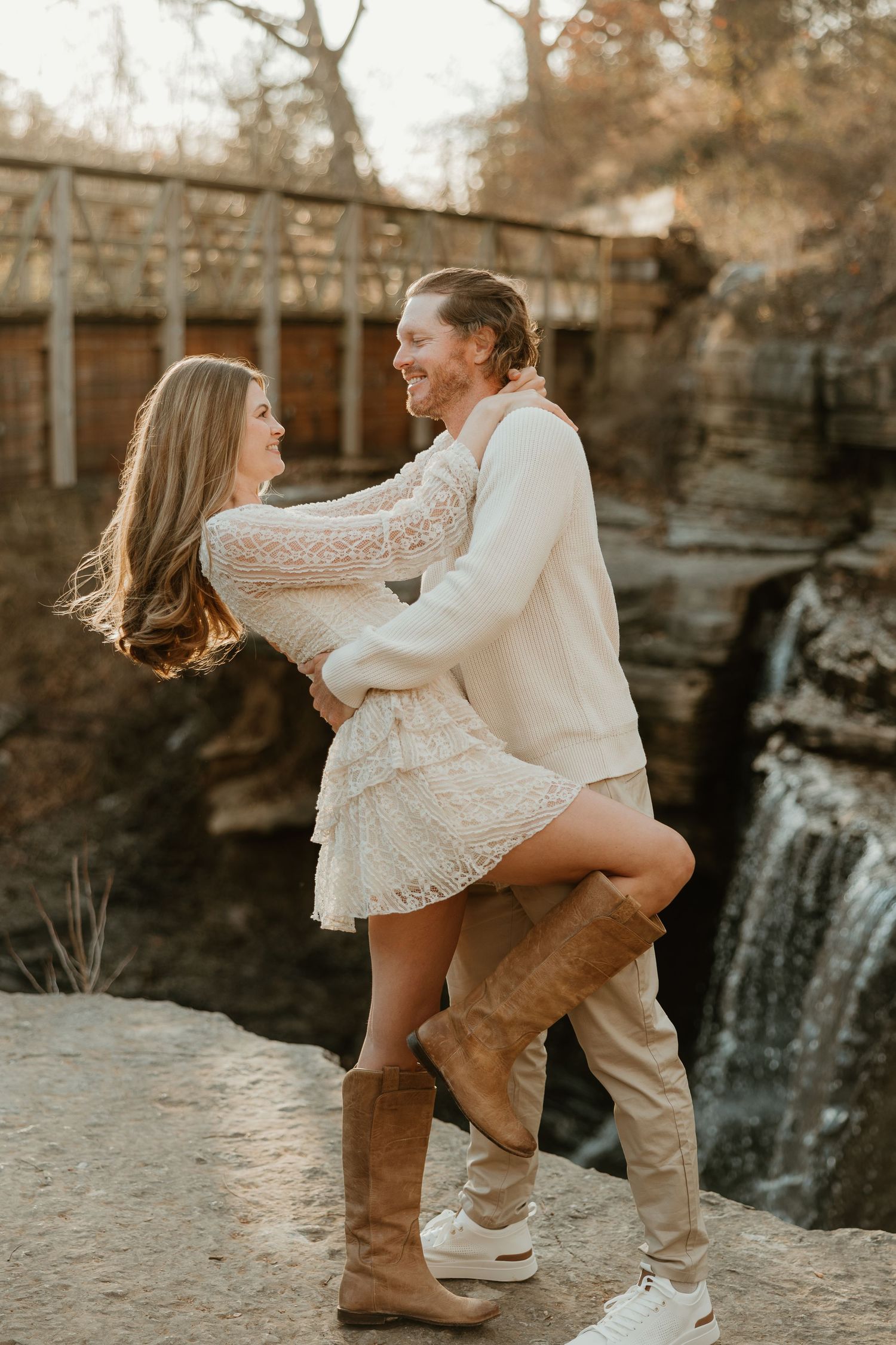 A couple embraces in a romantic moment at sunset near a rustic bridge and waterfall.