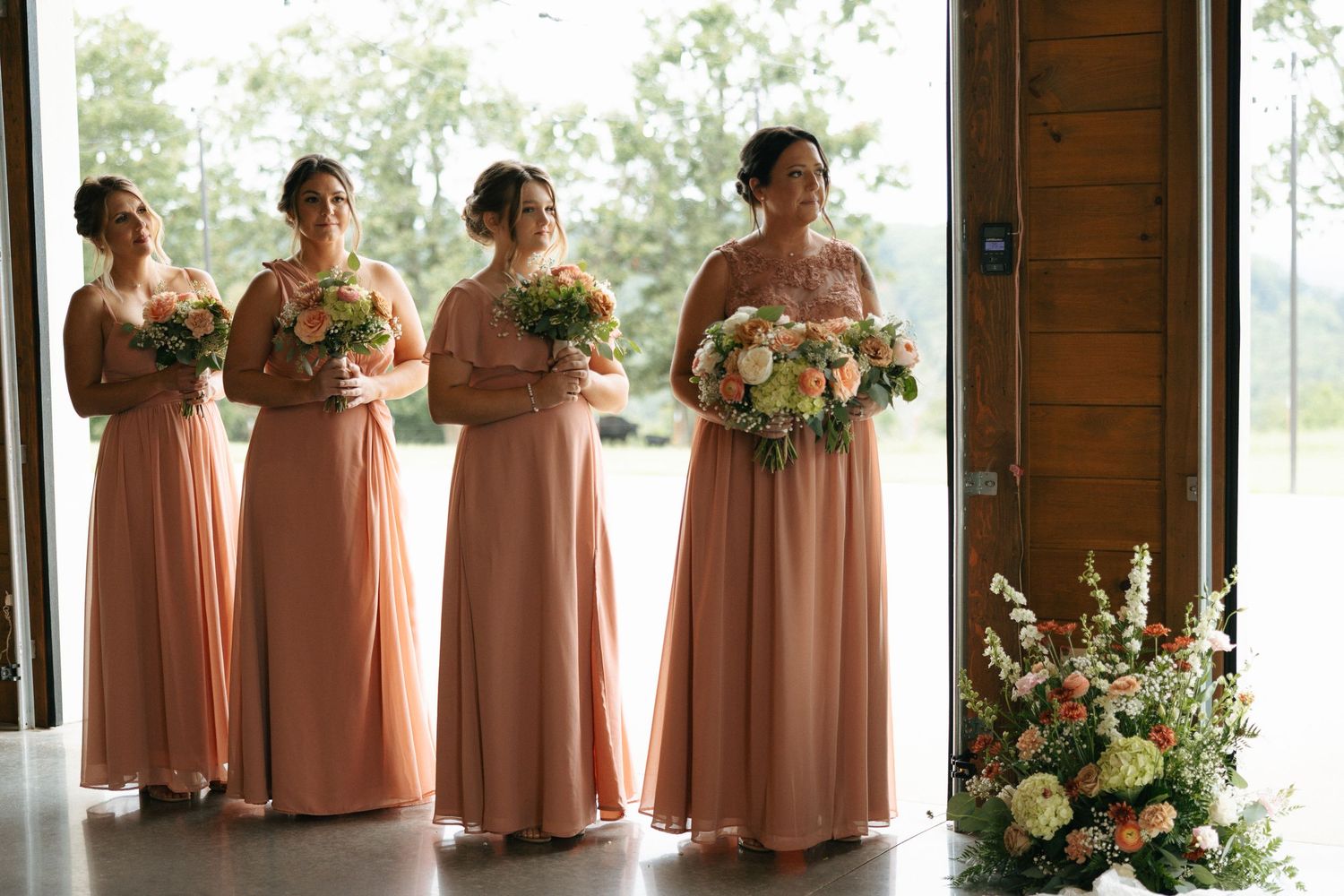 Bridesmaids in peach-colored dresses stand in a row at a rustic wooden venue decorated with floral arrangements.