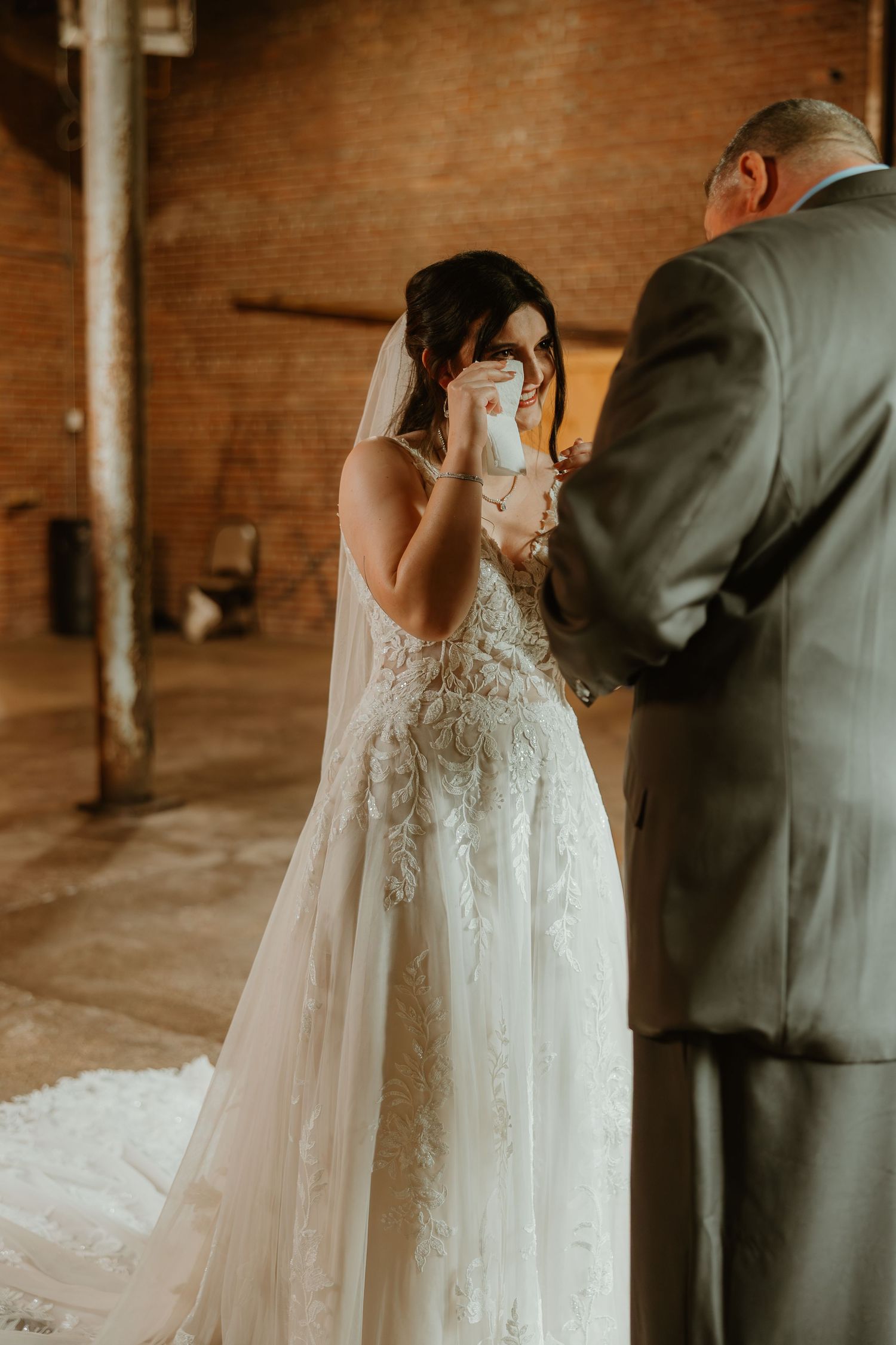 Emotional moment captured at indoor rustic brick venue as bride in lace gown wipes tears during ceremony.