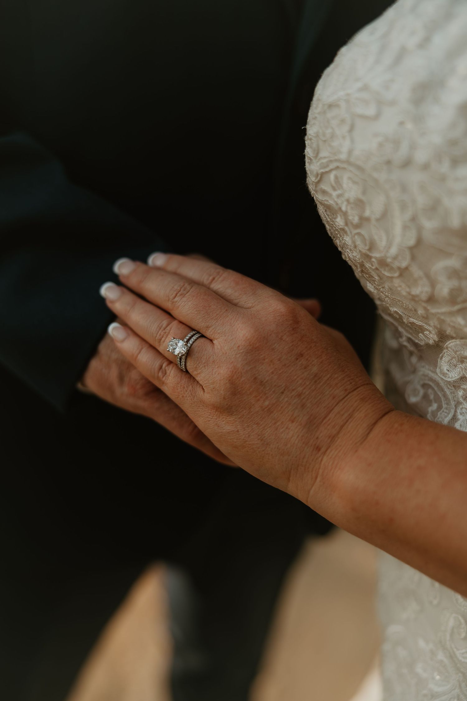 Close-up shot of wedding rings on hand showing white lace dress sleeve detail in moody natural lighting.