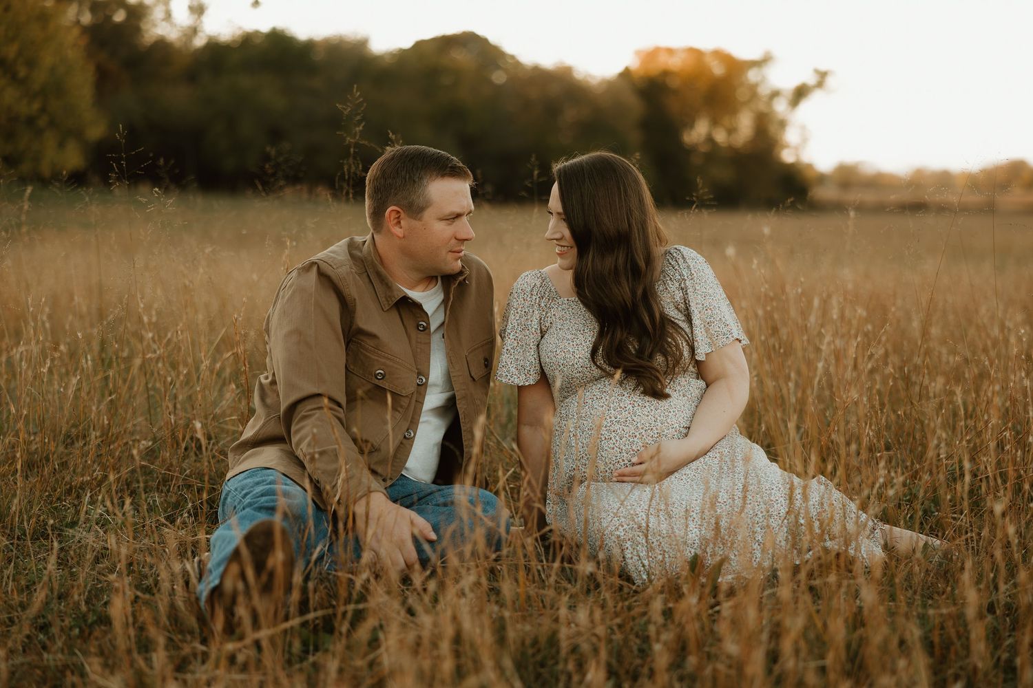 A couple sits intimately together in a golden wheat field during sunset, creating a romantic outdoor portrait.