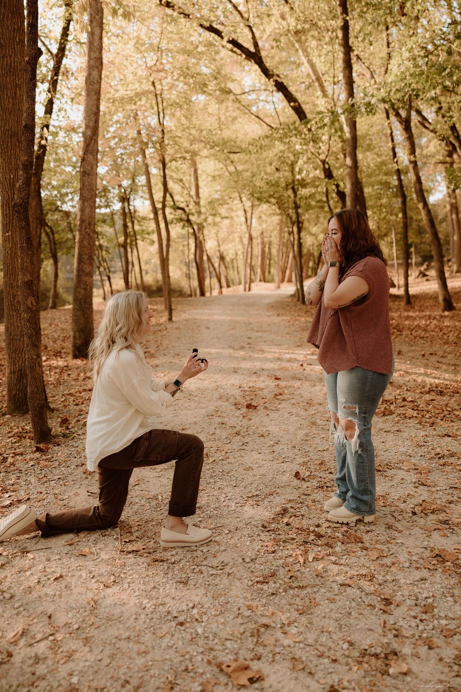 Two figures on a tree-lined autumn path, one kneeling while the other stands and captures the moment with a camera.