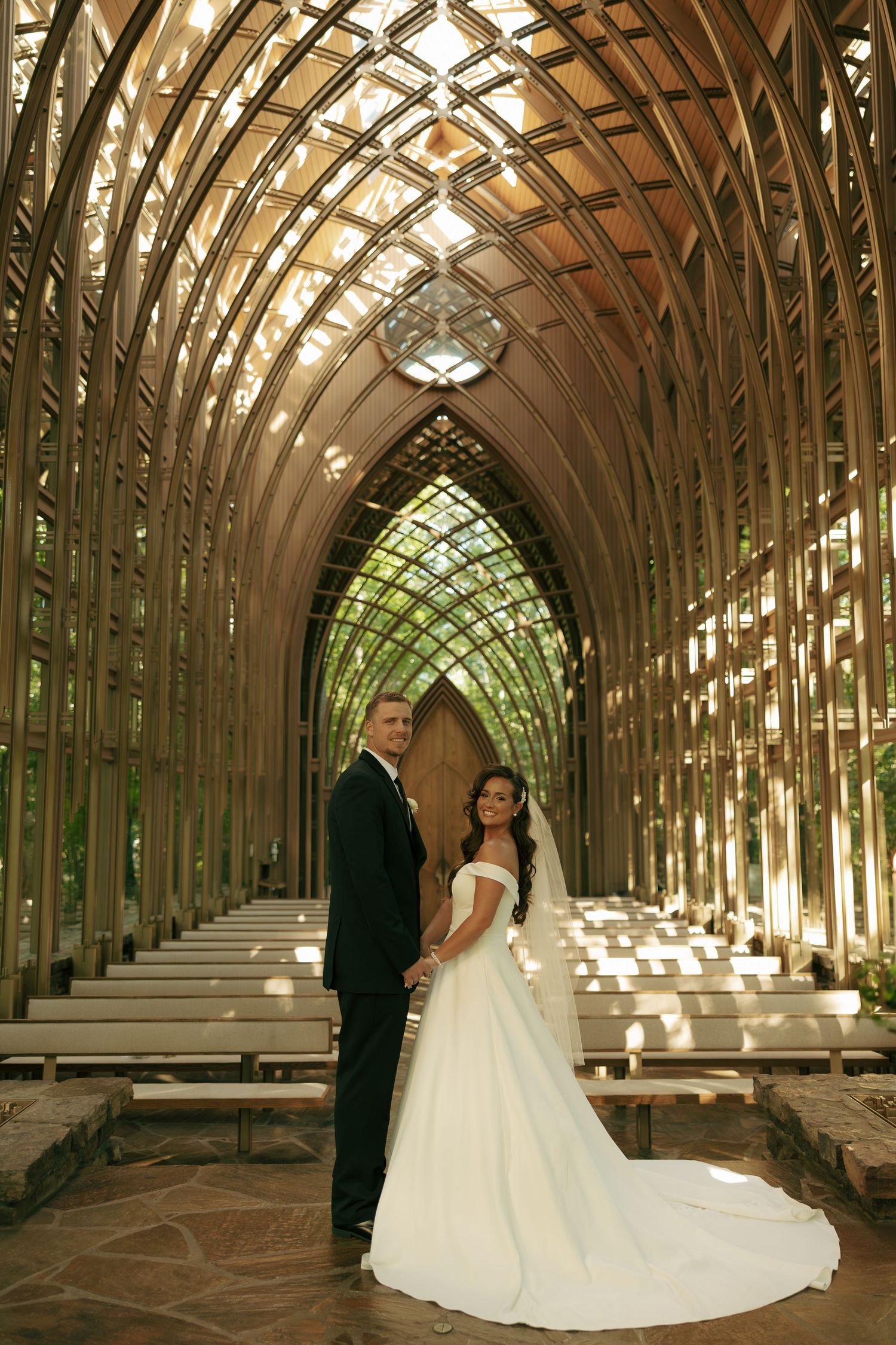 Bride and groom pose inside a stunning Gothic wood chapel with arched ceiling and natural light streaming through glass walls.