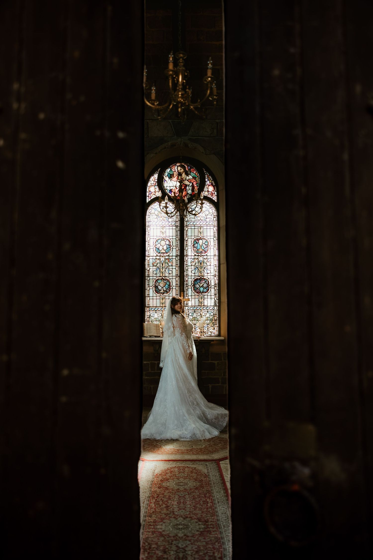 A wedding dress silhouetted against a stained glass window in a dark church creates a dramatic bridal portrait.