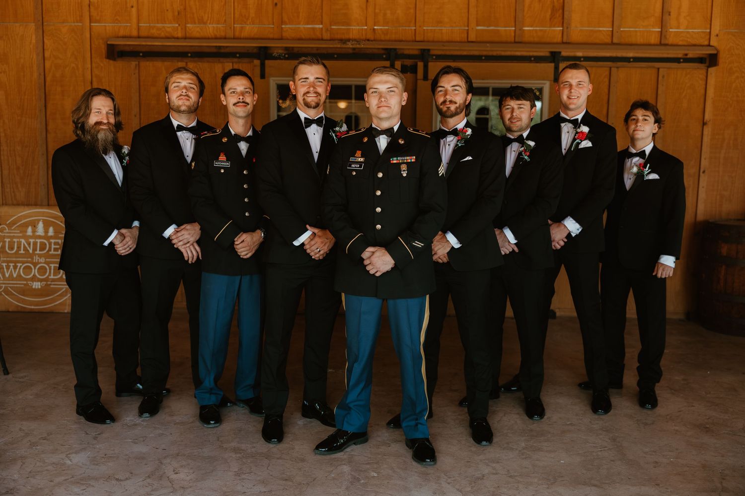 A group wearing formal black suits and blue jeans pose together for a wedding party photo in a rustic setting.