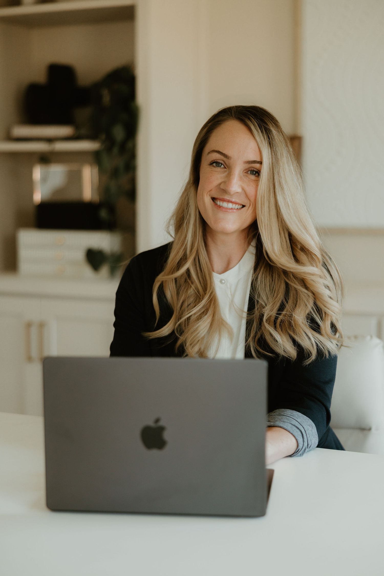 Professional working at white desk with MacBook laptop wearing navy blazer and white top in modern home office setting.