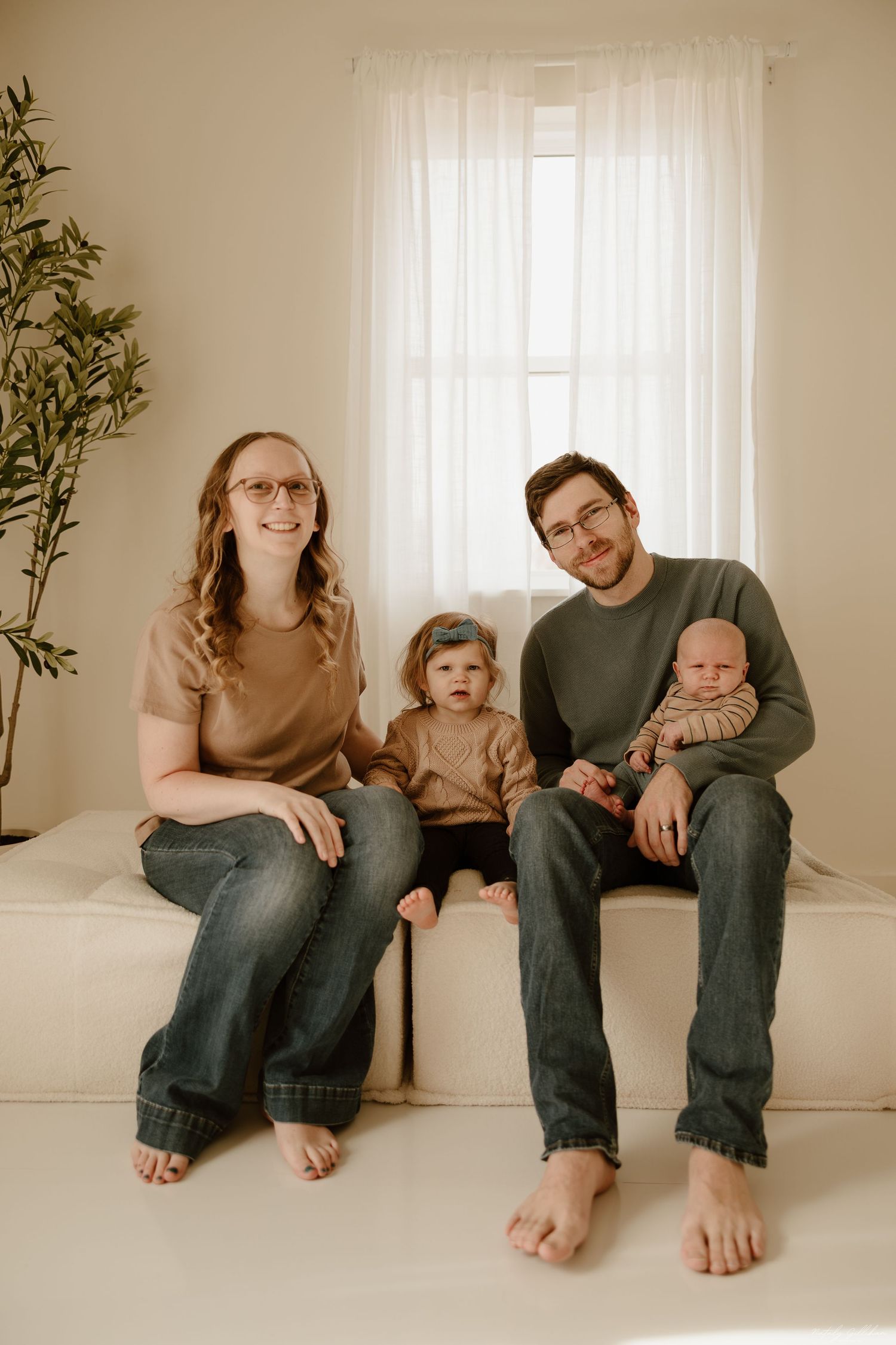A family of four sits barefoot on a white floor against a light wall with sheer curtains and a potted plant in the corner.