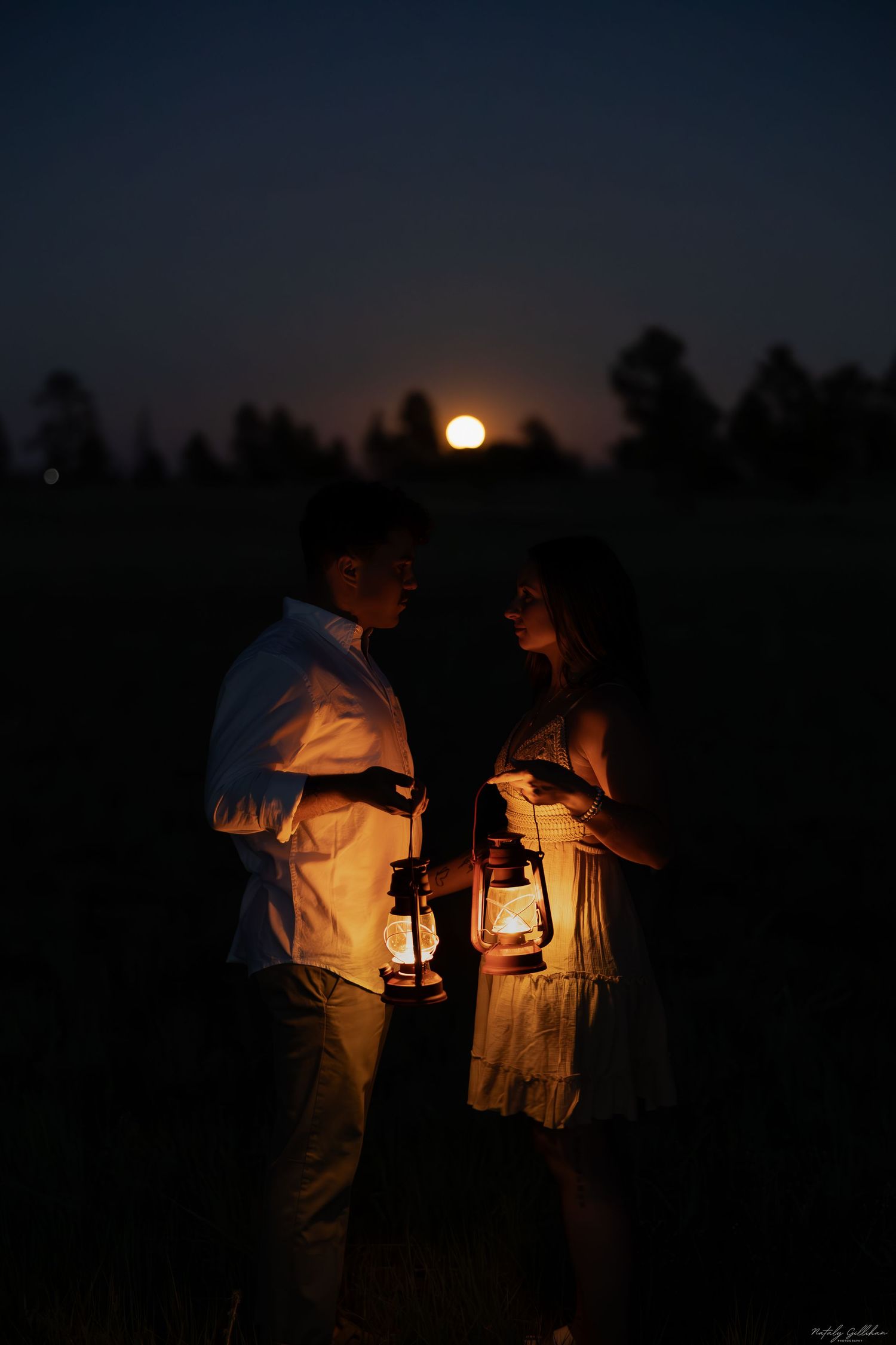 A couple holds lanterns at night with a full moon rising behind silhouetted trees in a romantic outdoor setting.