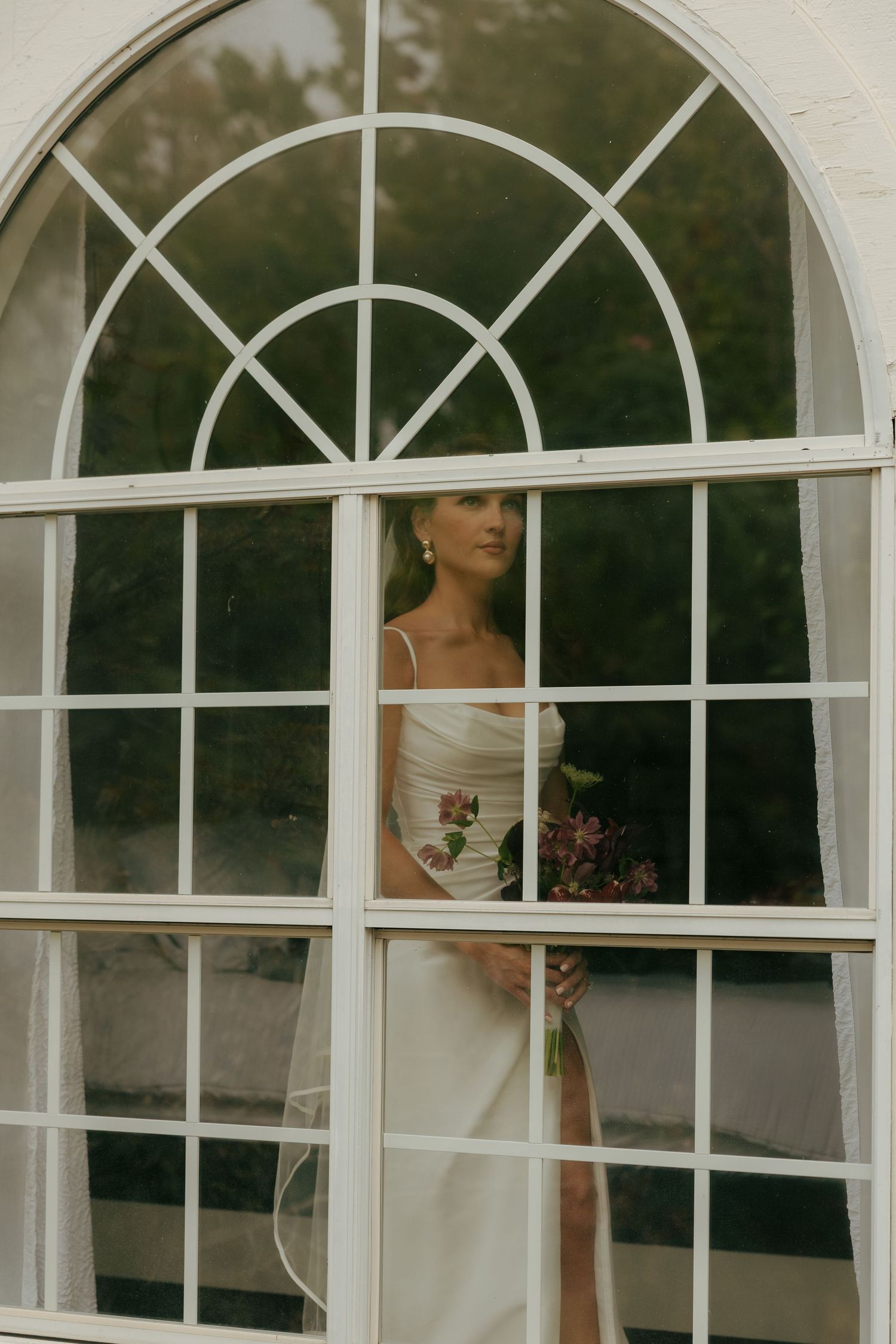 Bride in white gown holding floral bouquet, viewed through elegant arched window with white frames.