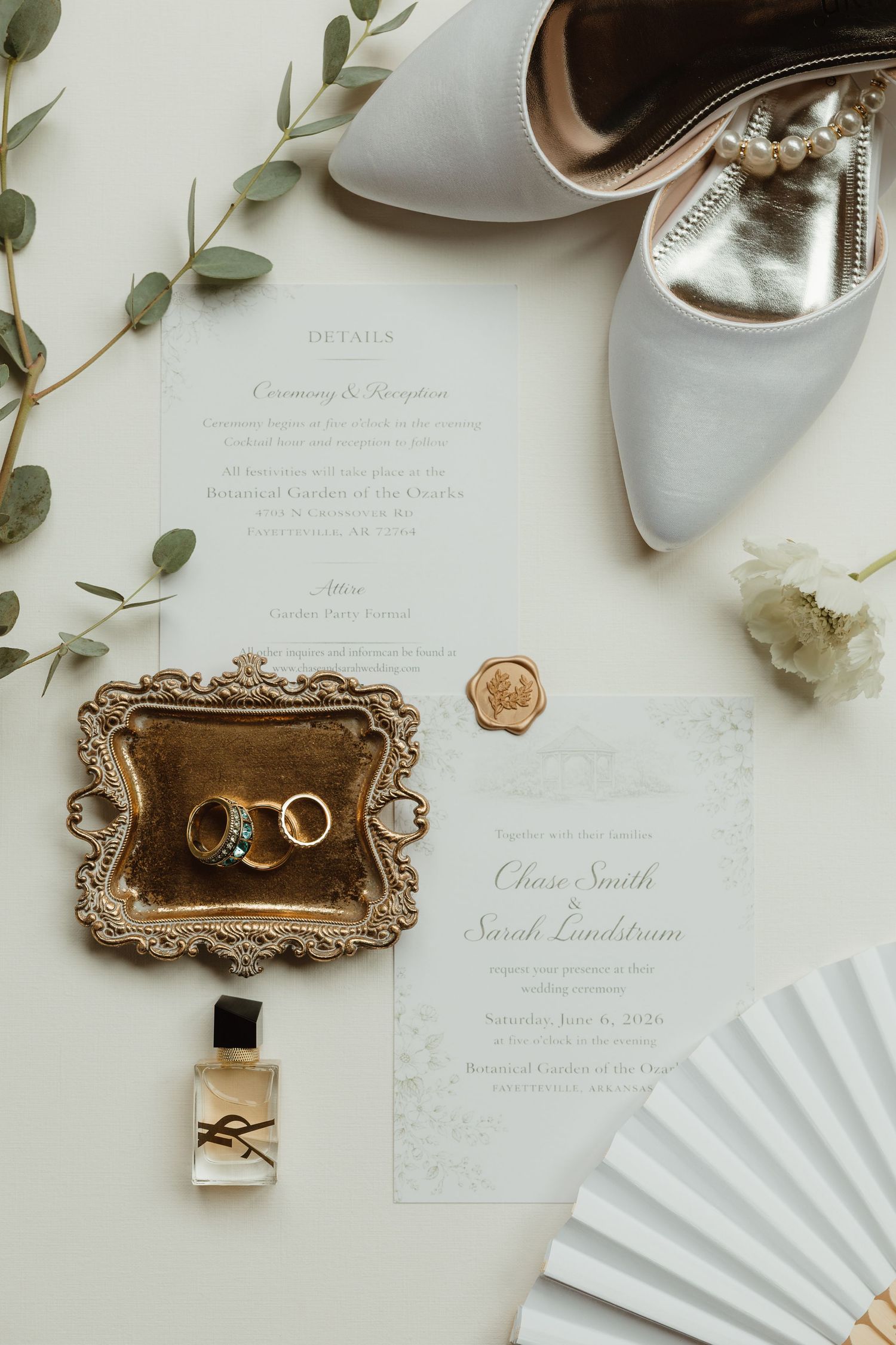 Elegant wedding flat lay with gold rings on ornate tray, white heels, YSL perfume, wax seal invitation, fan, and eucalyptus.