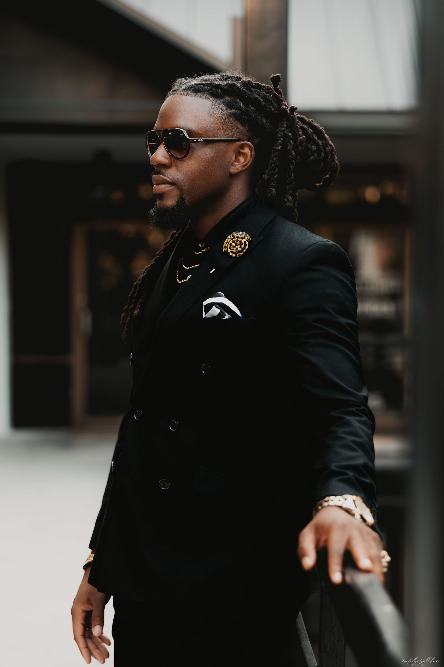 Stylish figure in a black blazer with dreadlocks and sunglasses standing against an urban backdrop.