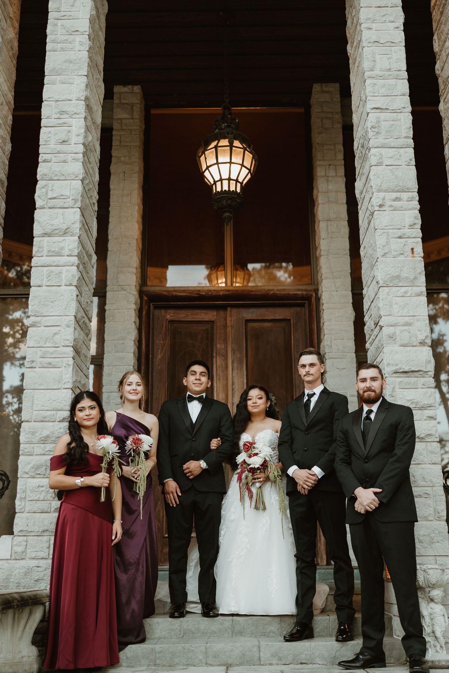 Wedding party stands in elegant formation against white stone columns at dusk with warm lighting.