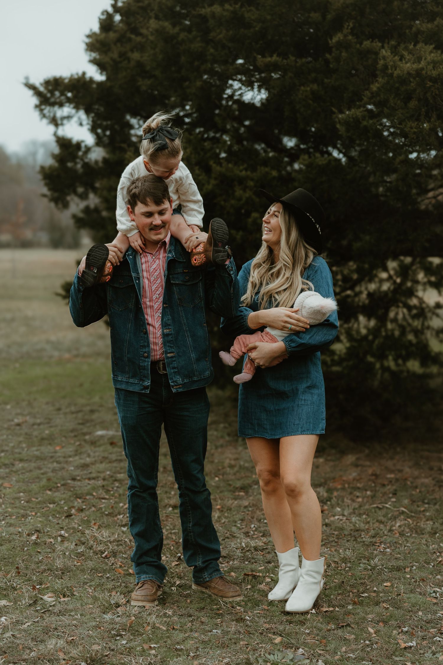 A young family poses outdoors, man carries toddler on shoulders while woman holds infant, both smiling in casual denim outfits.