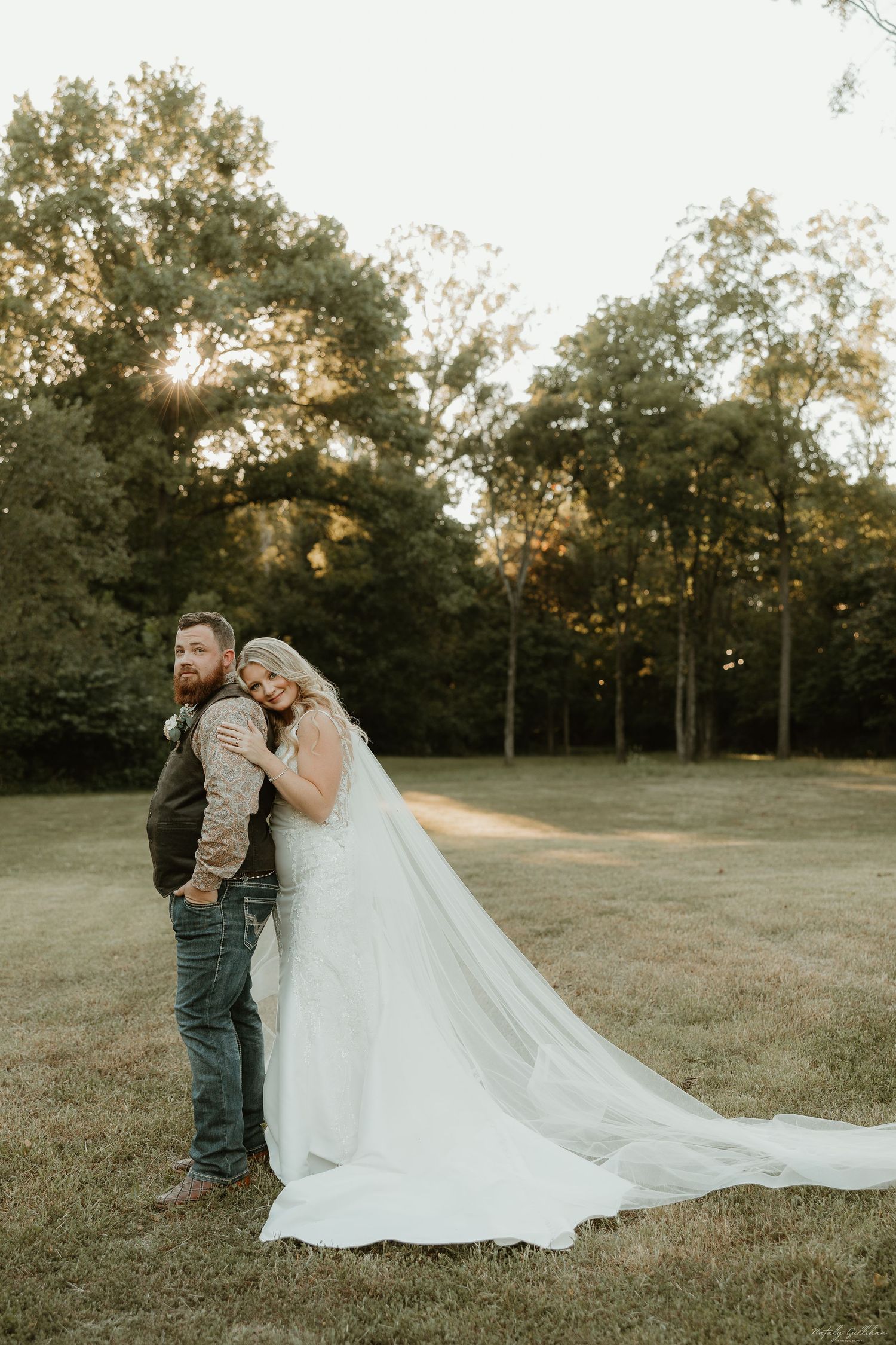 A couple in wedding attire stands back-to-back outdoors at sunset with a flowing white veil against a wooded backdrop.