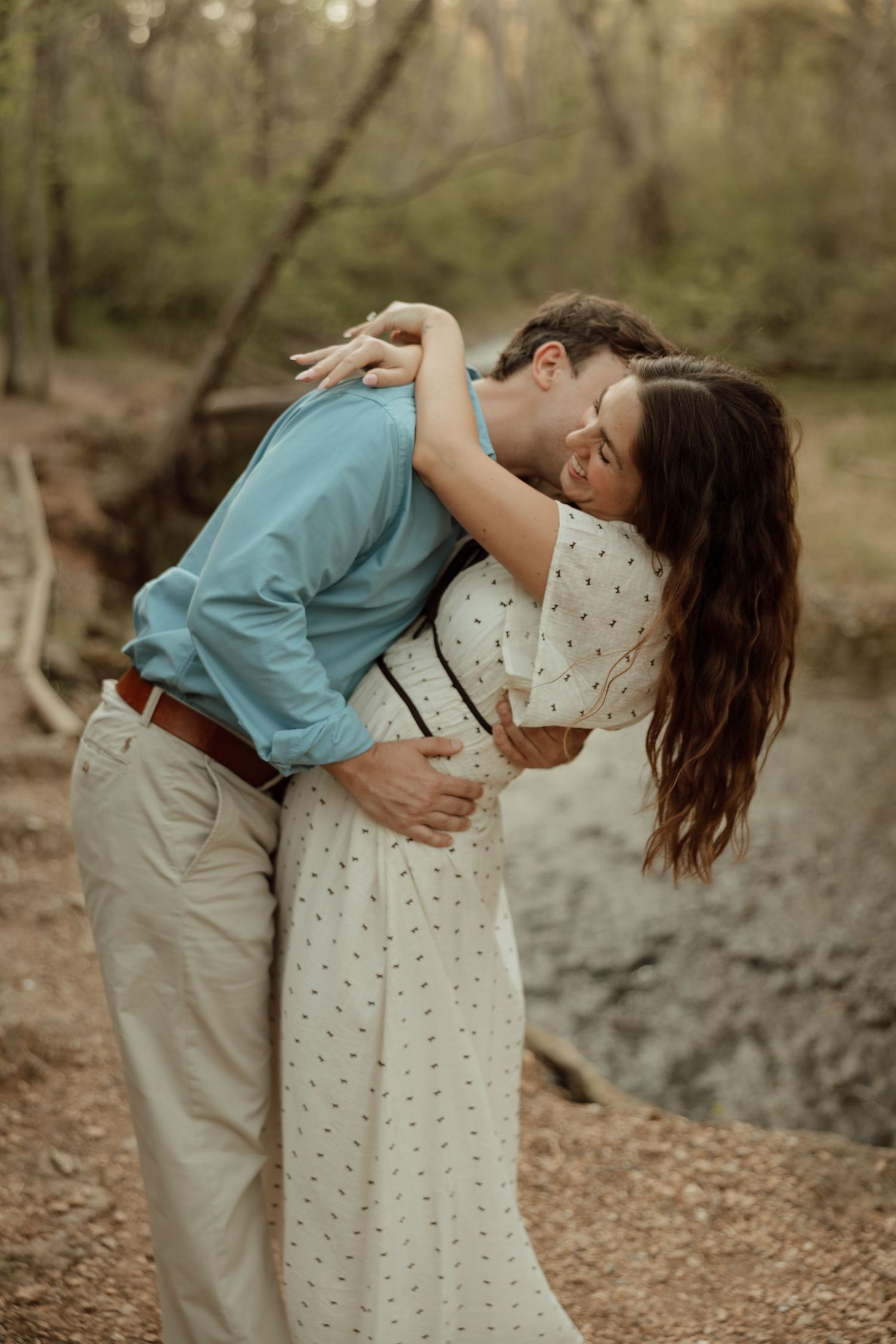 A couple shares a romantic embrace in a wooded outdoor setting, the man in blue kissing the woman in a white floral dress.