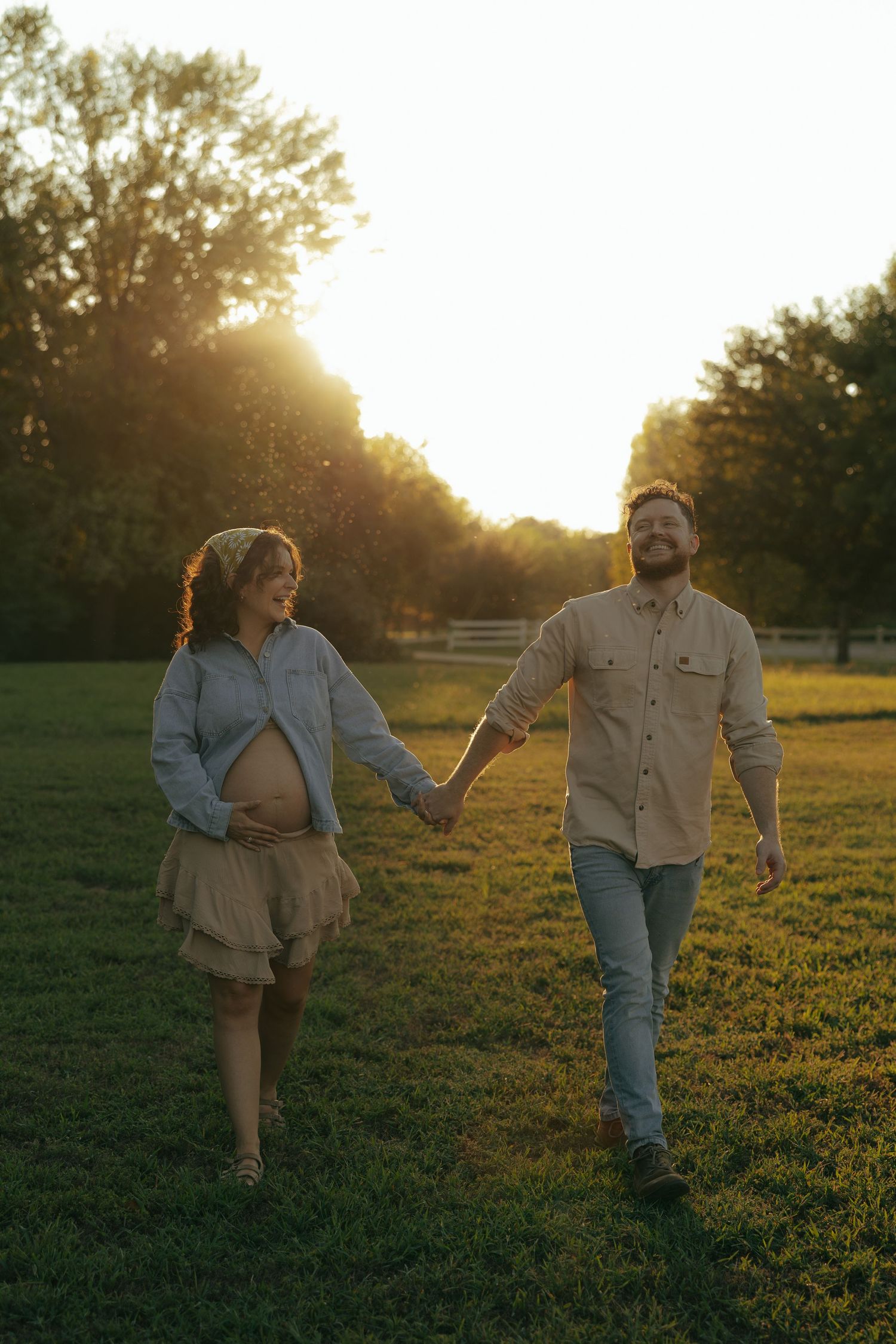 Pregnant couple holding hands walking through a sunlit green field at golden hour sunset.
