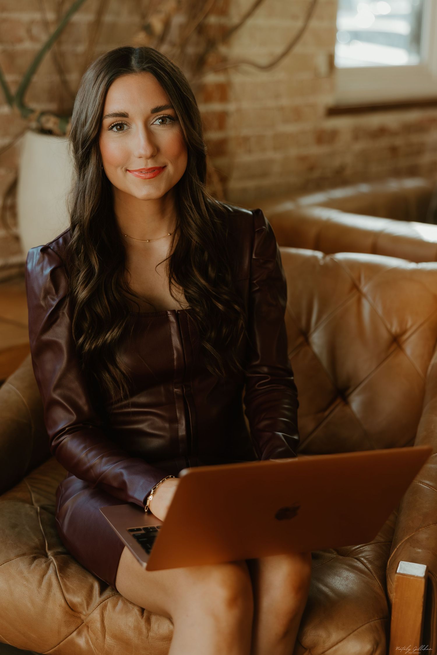 Professional working with laptop while sitting on brown leather couch in room with exposed brick wall.