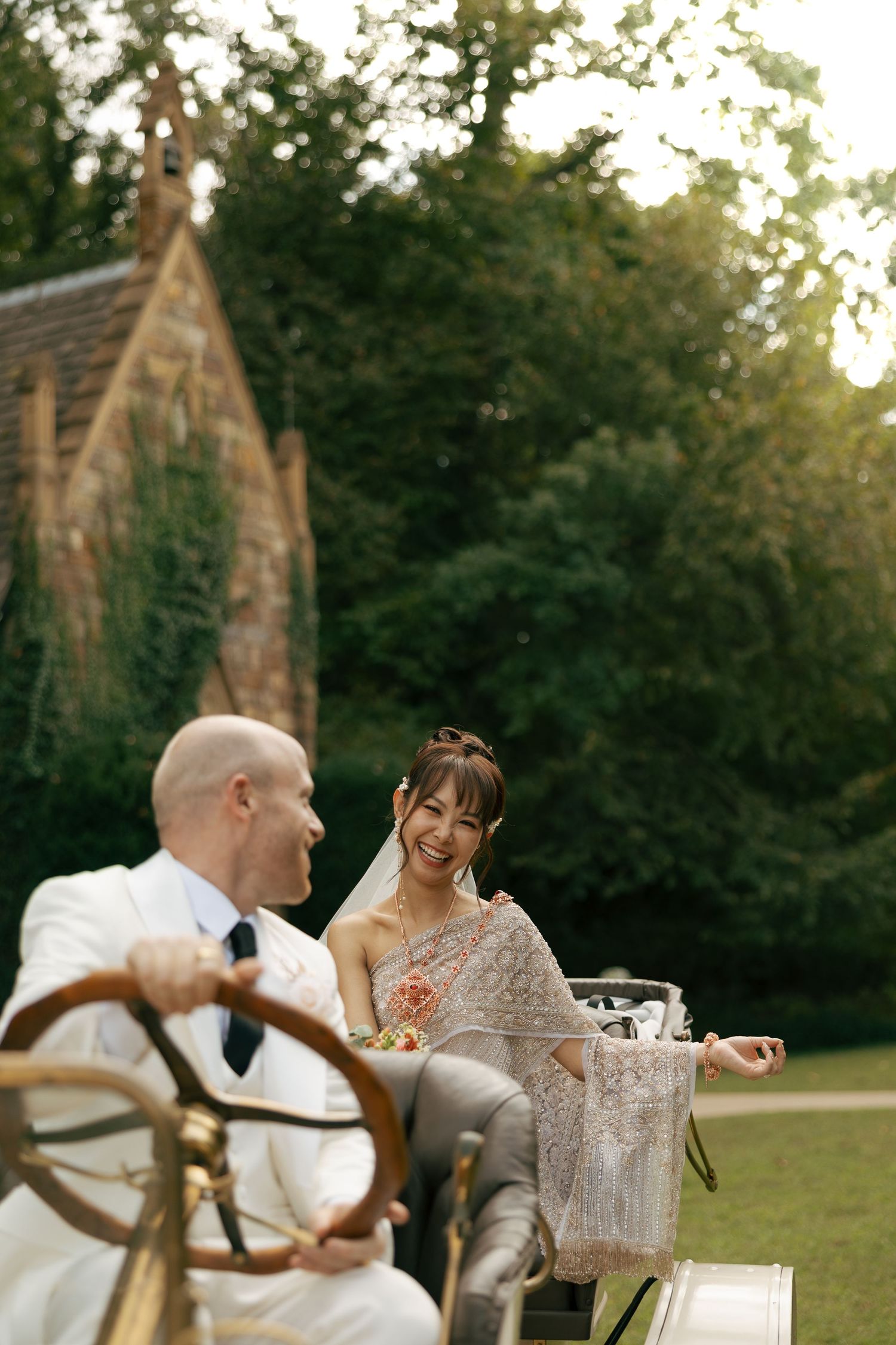 Bride and groom in vintage car at sunset, smiling, lush green trees and stone chapel in background.