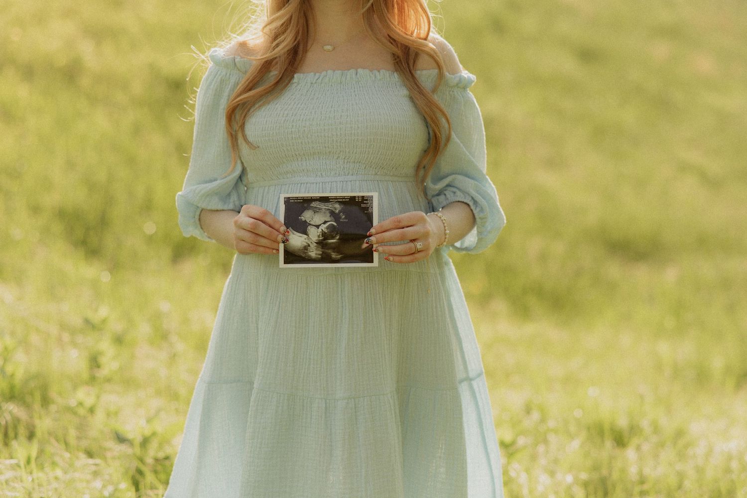 Someone in a flowing mint green dress holds an ultrasound photo while standing in a sunlit meadow.