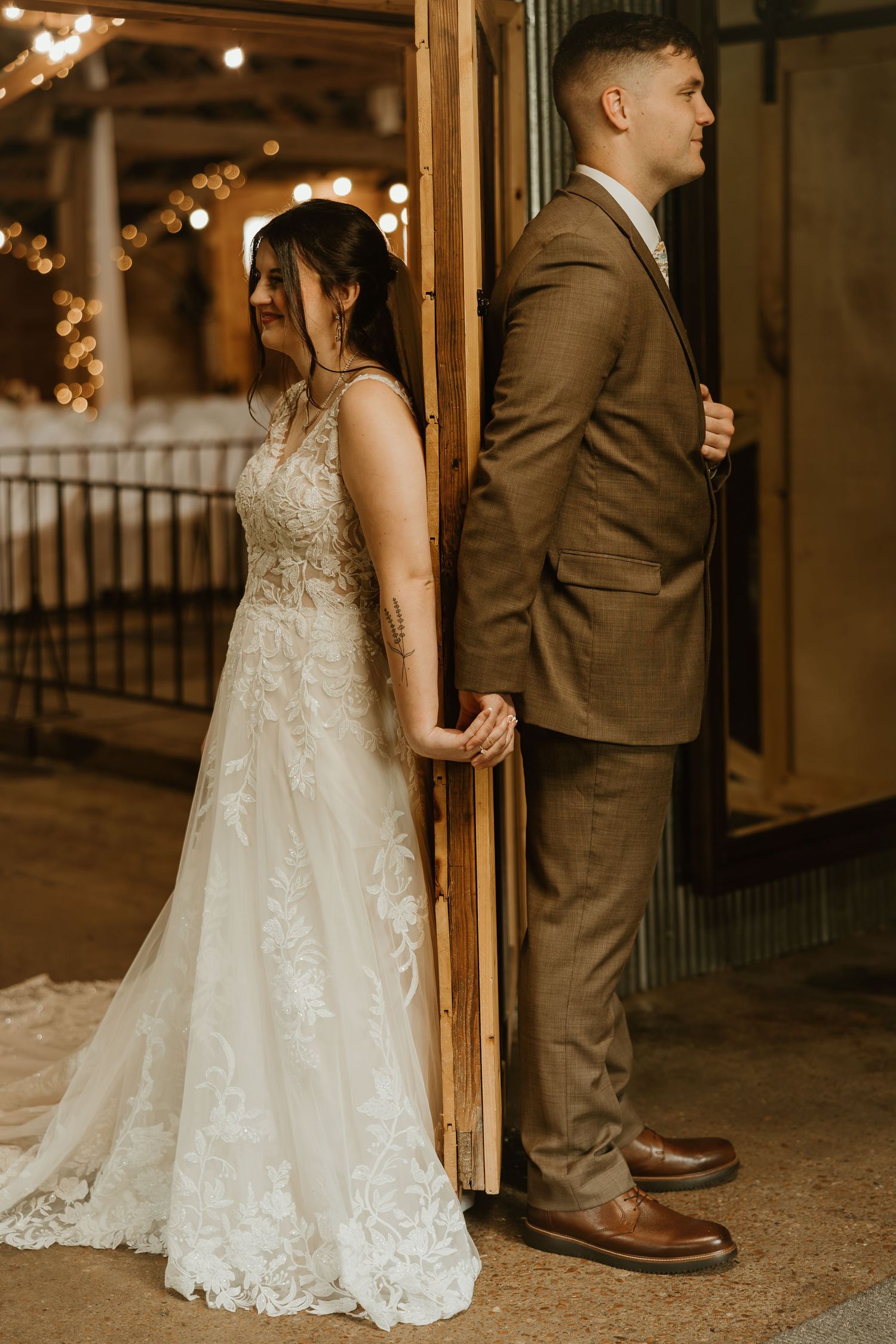 Couple stands back-to-back in formal wedding attire with bride in lace gown and groom in brown suit against twinkle lights.