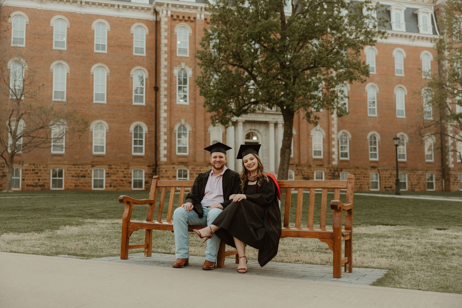 A group of people gather around a wooden bench on a college campus with historic brick buildings in the background.