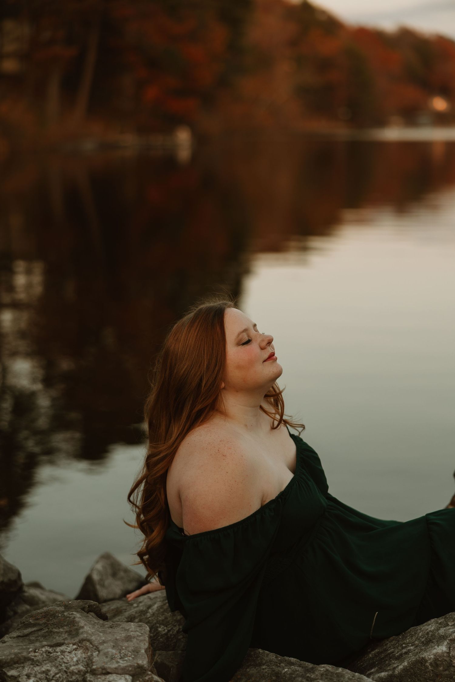 A woman in a black dress reclines on rocks by a tranquil lake during golden hour with autumn foliage reflections.